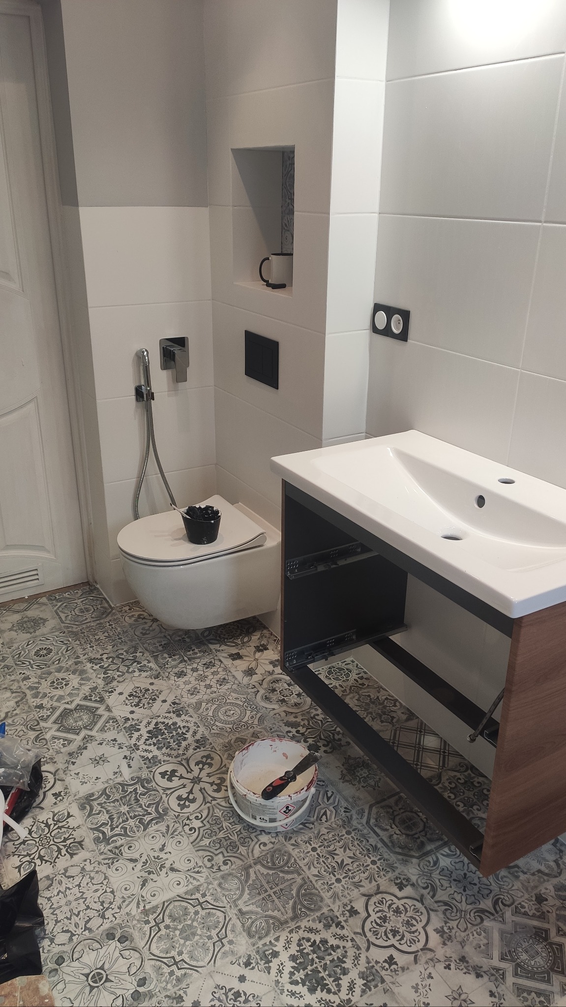 Montaż szafki umywalkowej w łazience z wzorzystą podłogą, widoczny biały sedes z bidetką i czarnymi akcesoriami, wiadro z zaprawą na podłodze.