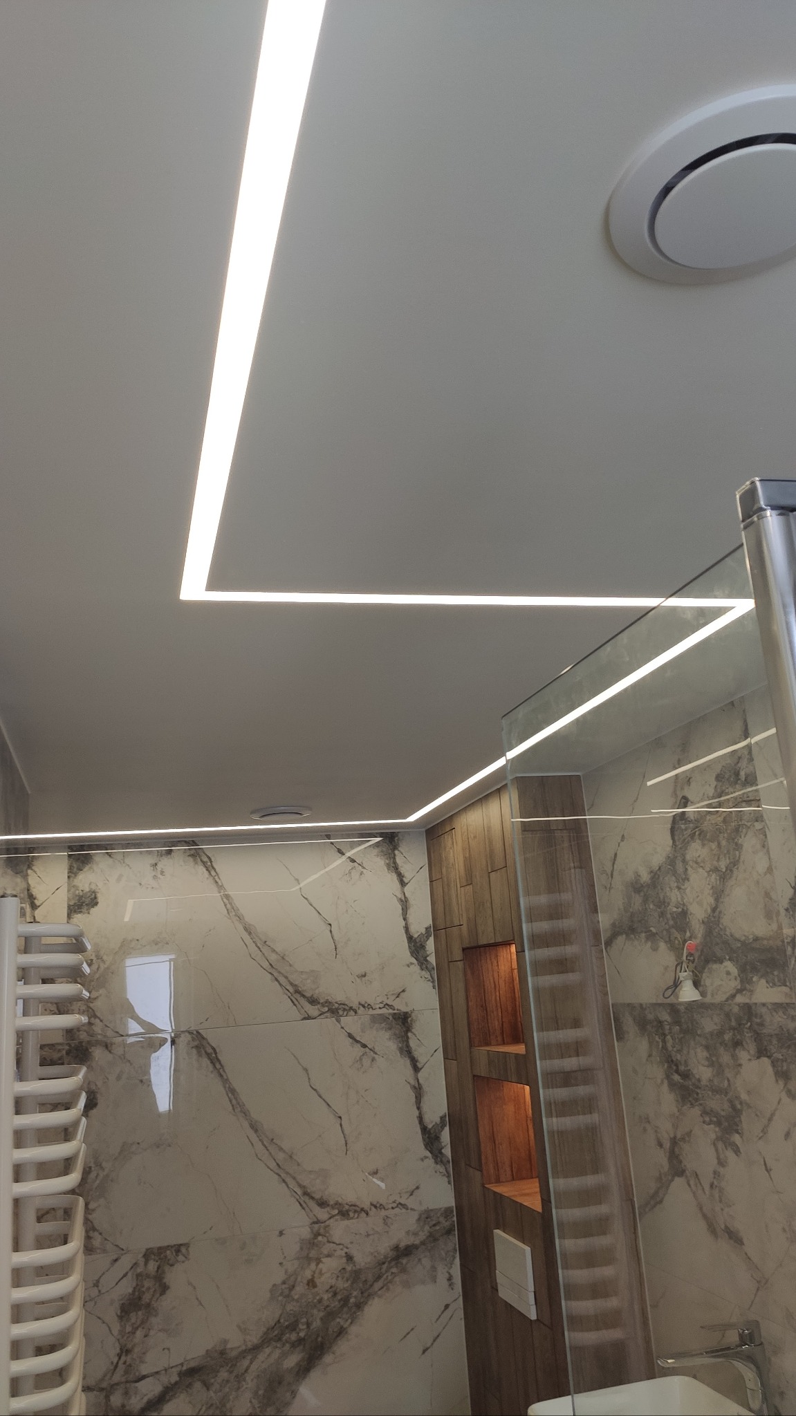 Nowoczesna łazienka w Jaśle z podświetlanym sufitem i marmurowymi płytkami na ścianach. Widoczny grzejnik drabinkowy i zabudowa z półkami.