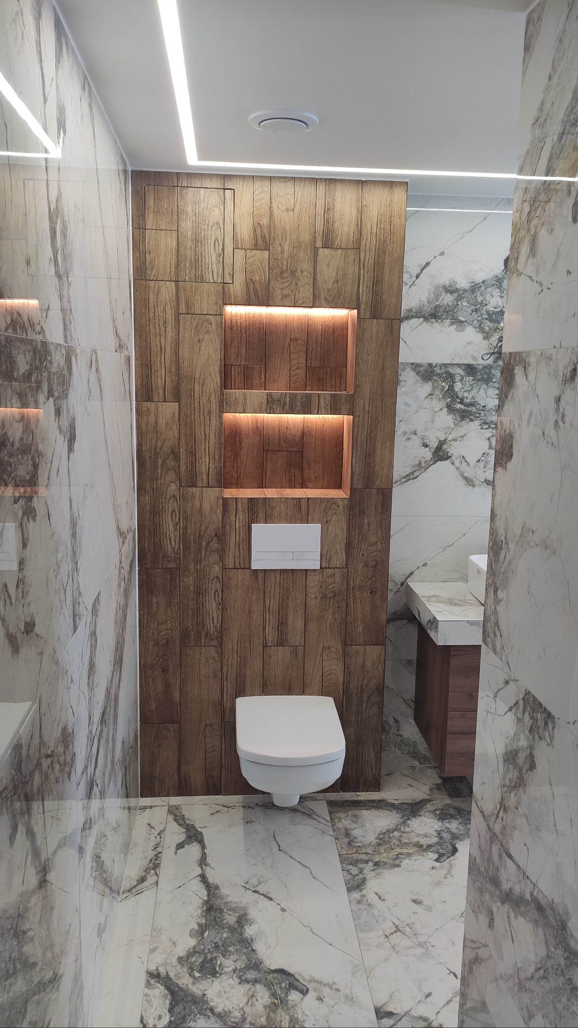 Nowoczesna łazienka z drewnianą ścianą akcentującą, oświetlonymi niszami i marmurowymi płytkami podłogowymi oraz wiszącą miską WC.