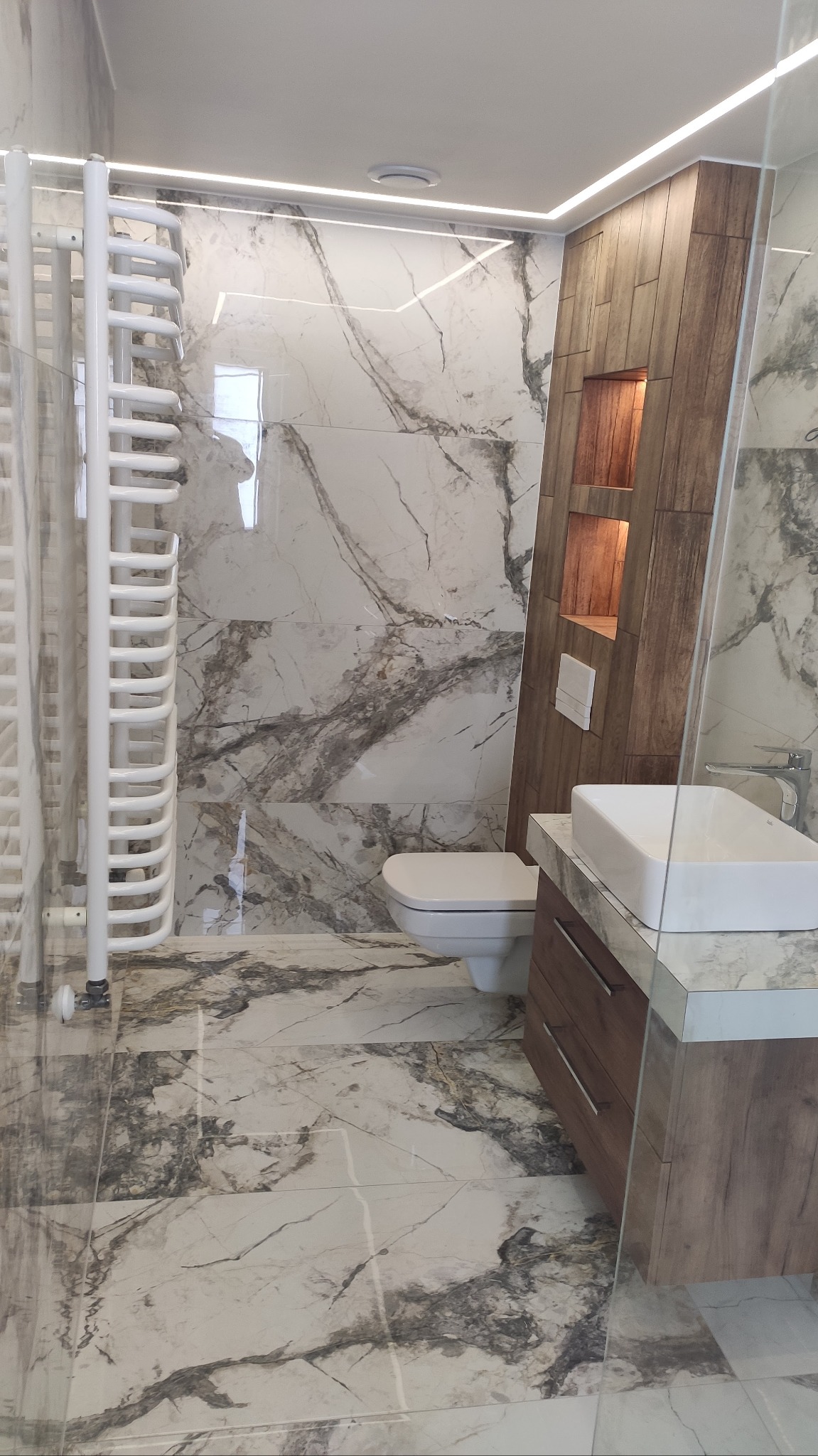 Nowoczesna łazienka z marmurowymi płytkami, podświetlanym sufitem, grzejnikiem drabinkowym i drewnianym elementem zabudowy z półkami.