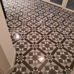 Wzorzysta podłoga z płytek ceramicznych w odcieniach szarości i bieli, ułożona w geometryczny, symetryczny deseń, widoczna w częściowo oświetlonym pomieszczeniu z białymi ścianami i drzwiami...