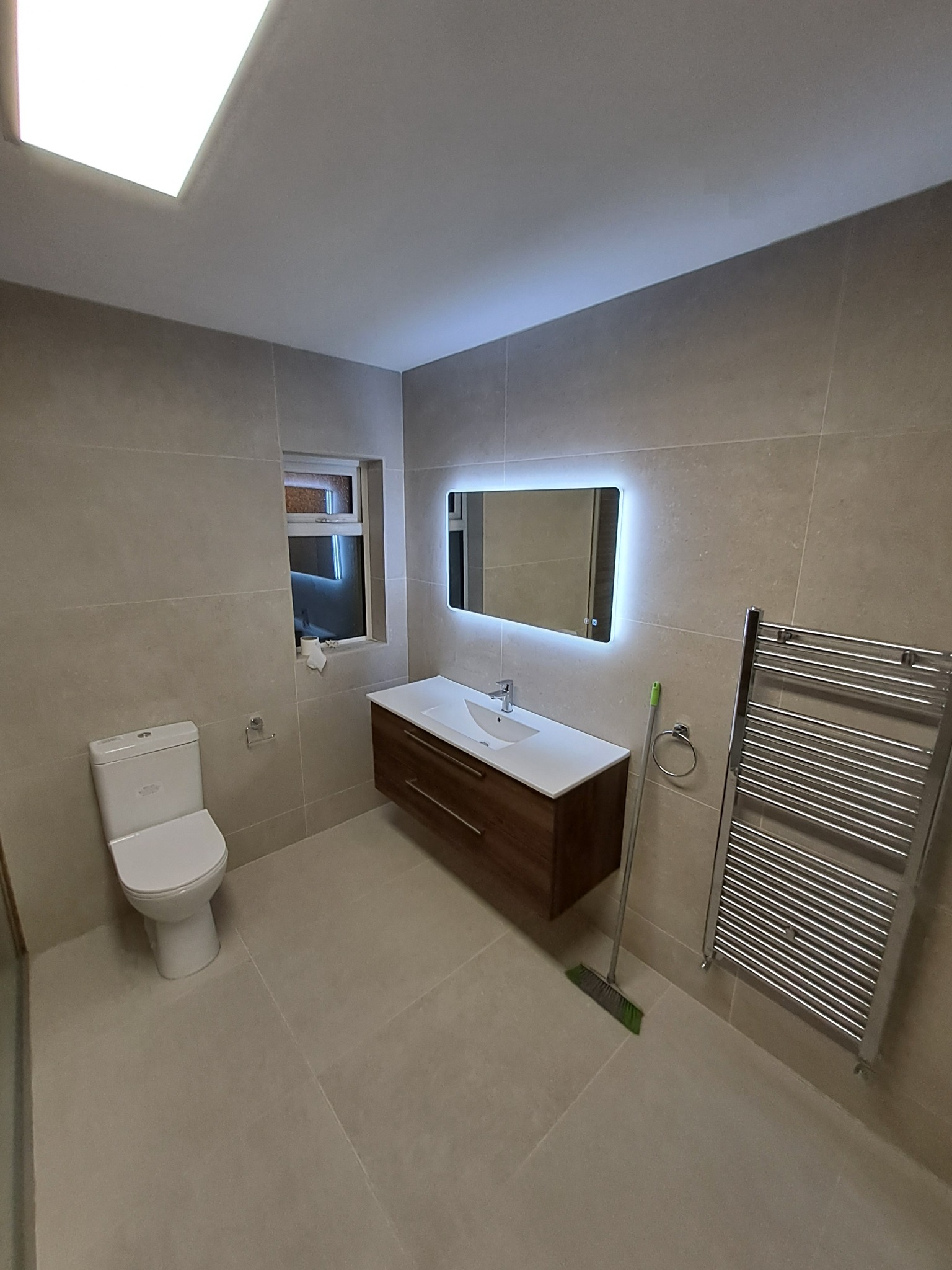 Nowoczesna łazienka z beżowymi płytkami, podświetlanym lustrem nad umywalką z szafką w kolorze ciemnego drewna, wiszącą toaletą i grzejnikiem drabinkowym. Na podłodze stoi zielona zmiotka.