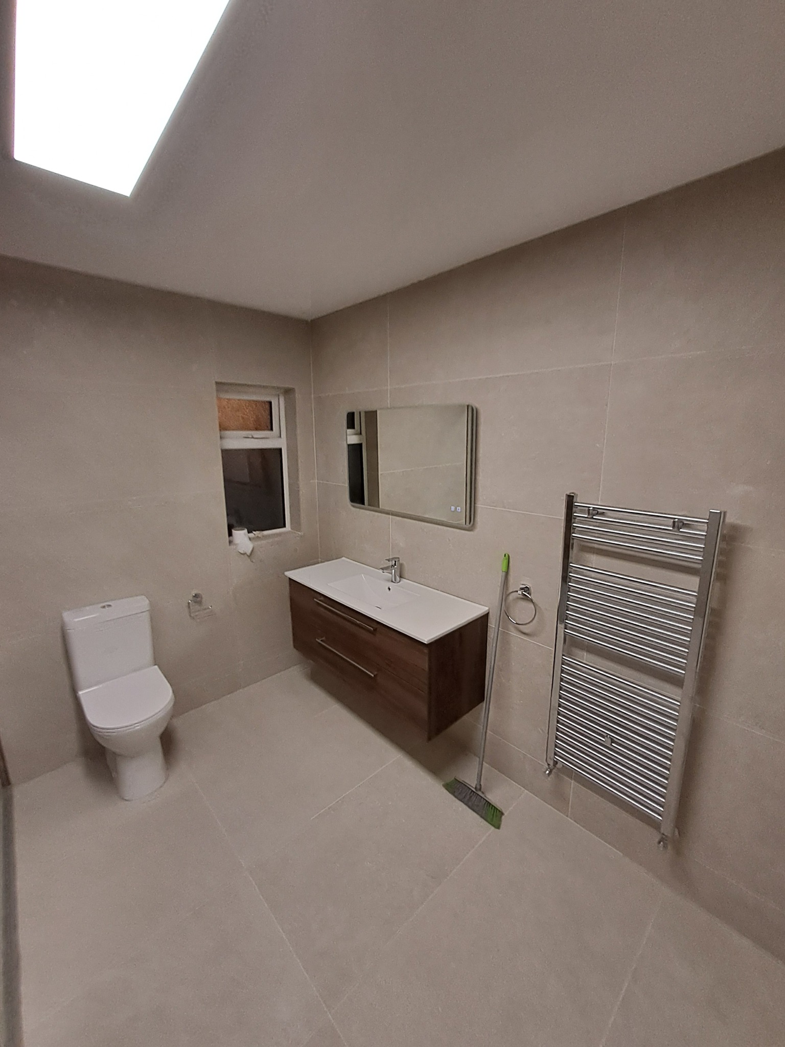 Nowoczesna łazienka po remoncie z beżowymi płytkami, podwieszaną szafką z umywalką, grzejnikiem drabinkowym i oknem. Użyto mopa do posprzątania.