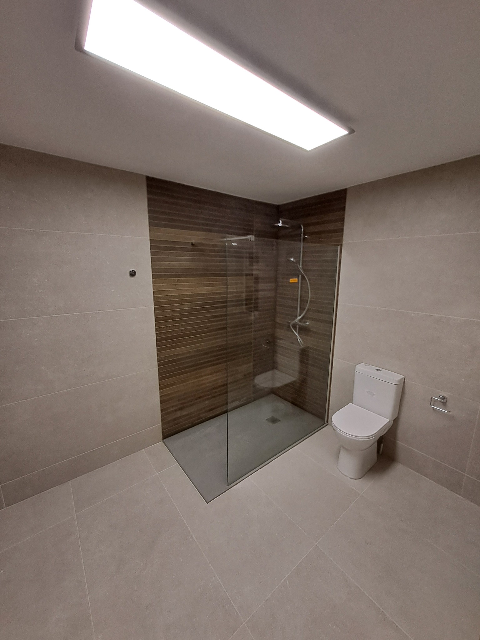 Nowoczesna łazienka z kabiną prysznicową typu walk-in, wykończona płytkami w odcieniach beżu i brązu, z widoczną toaletą i oświetleniem sufitowym.