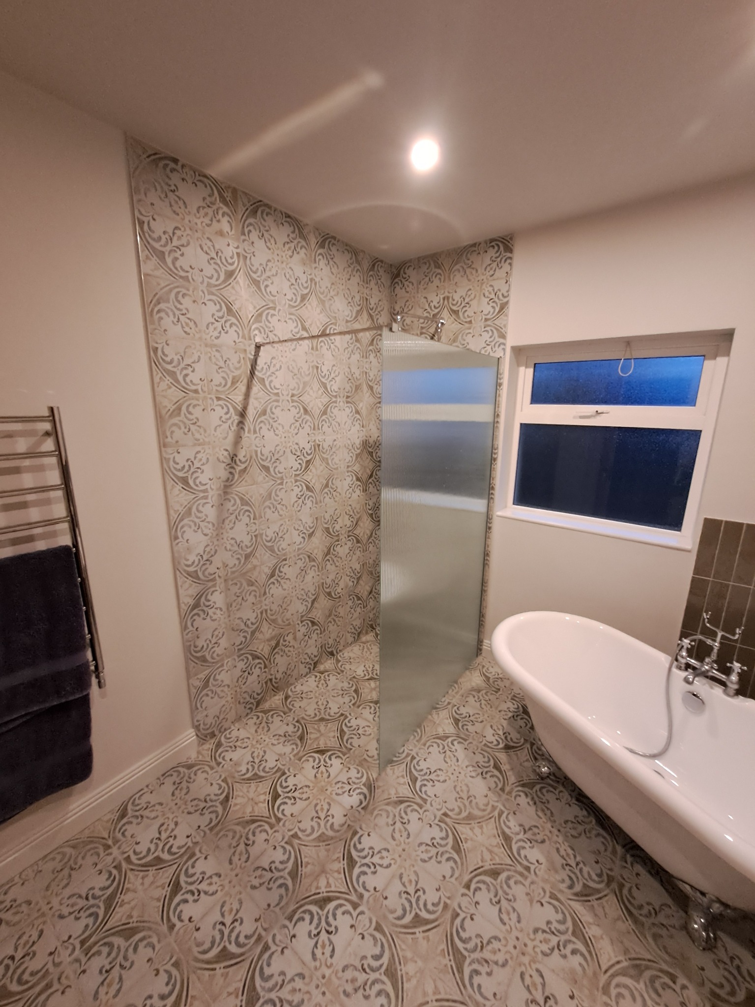 Wykończona łazienka z kabiną prysznicową ze szklaną ścianką, wolnostojącą białą wanną na ozdobnych nóżkach i grzejnikiem drabinkowym z powieszonym ciemnym ręcznikiem, całość utrzymana w jasnej...