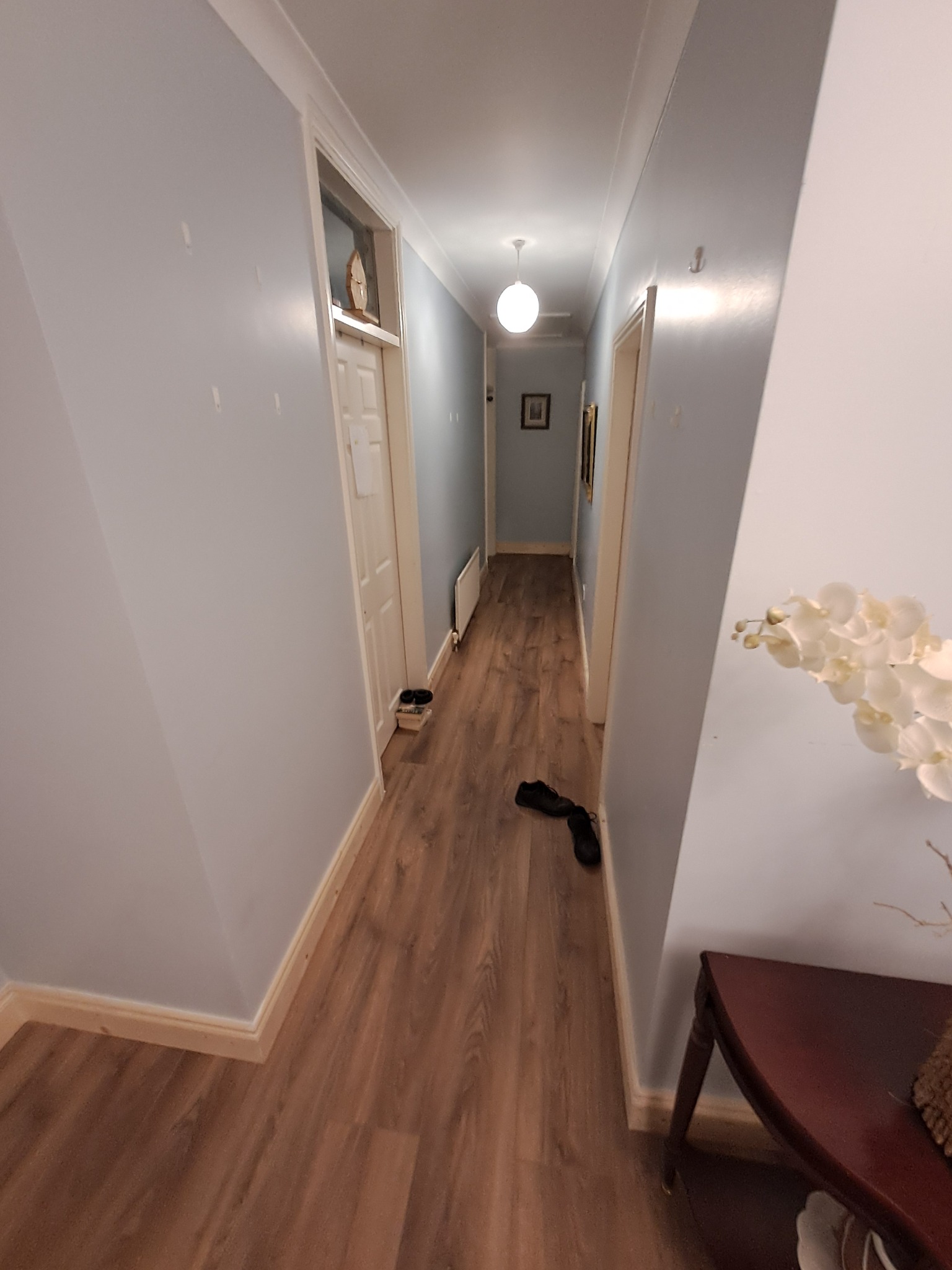 Długi korytarz w odcieniach szarości i brązu z panelami podłogowymi, widoczne drzwi, lampa sufitowa oraz dekoracyjny stolik z kwiatami po prawej stronie