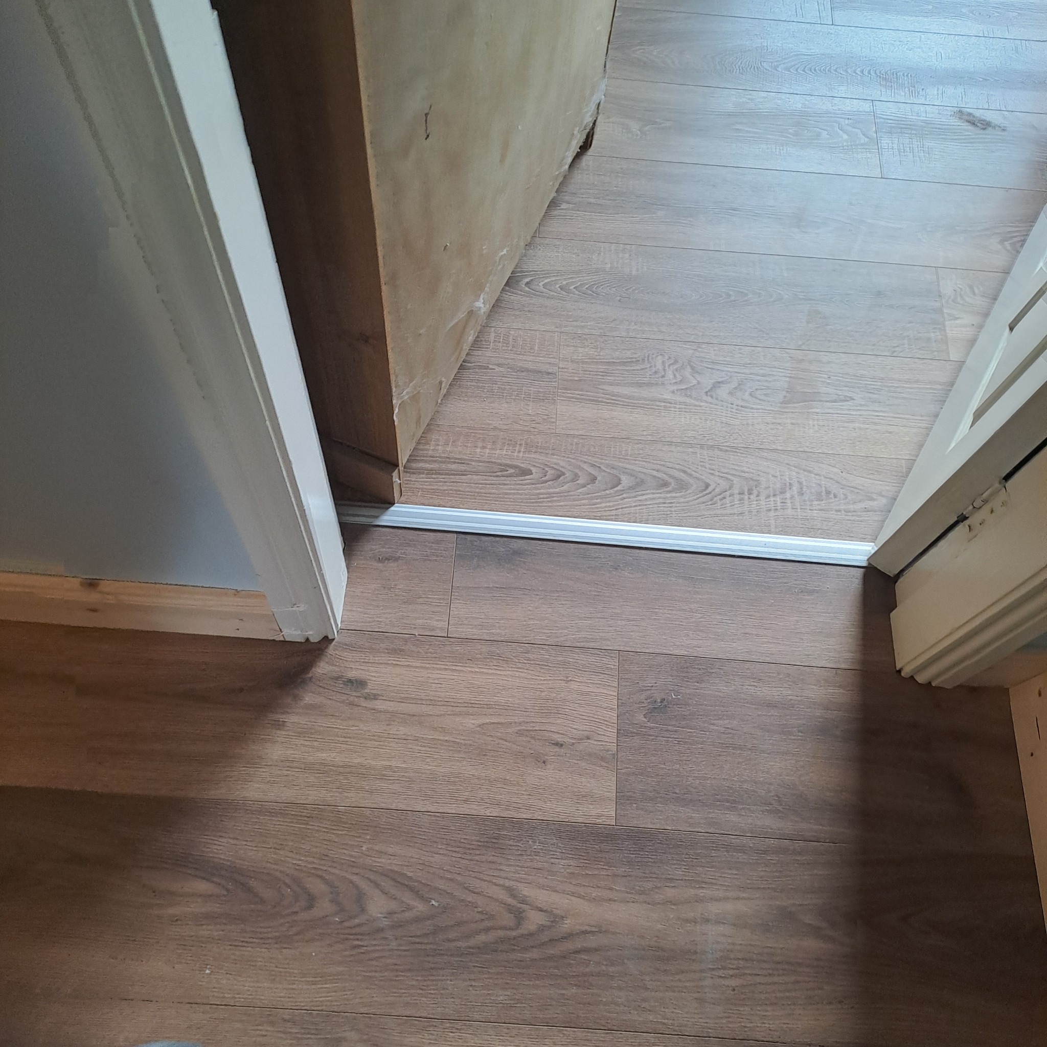 Ułożone panele podłogowe w odcieniach brązu, widoczna listwa progowa między pomieszczeniami, fragment ościeżnicy drzwi po prawej stronie.