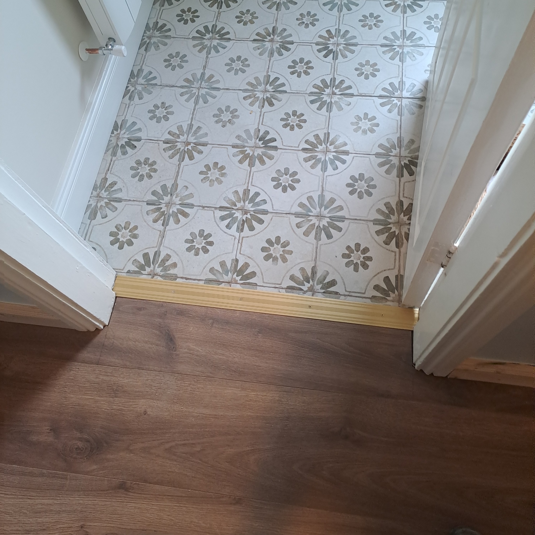 Połączenie dwóch rodzajów podłóg: brązowe panele podłogowe i płytki podłogowe w kwiatowy wzór, oddzielone złotą listwą progową, widok z góry na otwarte drzwi.