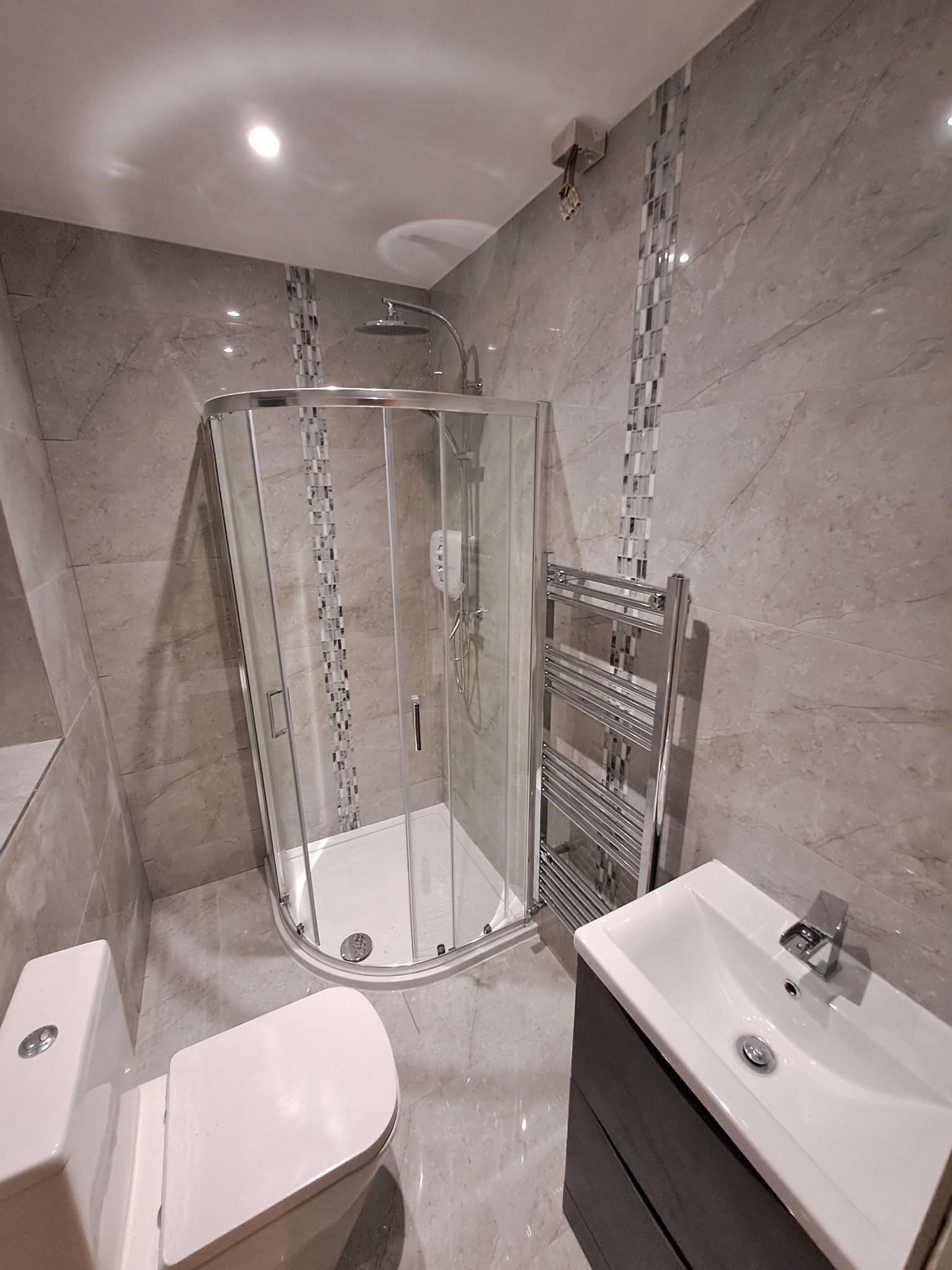 Kompletna łazienka w szarych odcieniach z kabiną prysznicową narożną, wiszącym grzejnikiem drabinkowym, umywalką z szafką i toaletą.