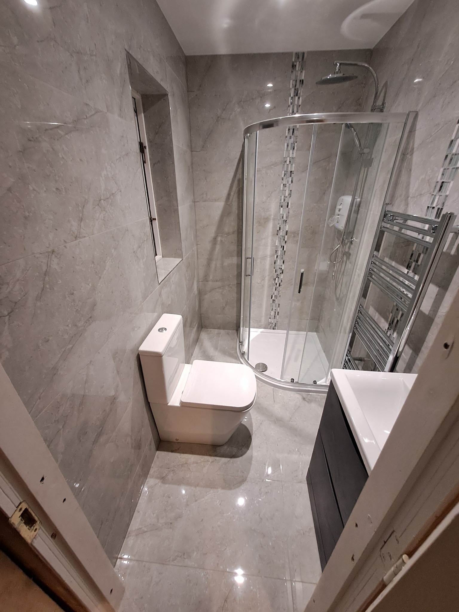 Wykończona łazienka z szarymi płytkami imitującymi marmur, kabiną prysznicową z chromowanymi detalami, podwieszaną toaletą i umywalką z szafką w kolorze antracytowym, widok zza otwartych drzwi.