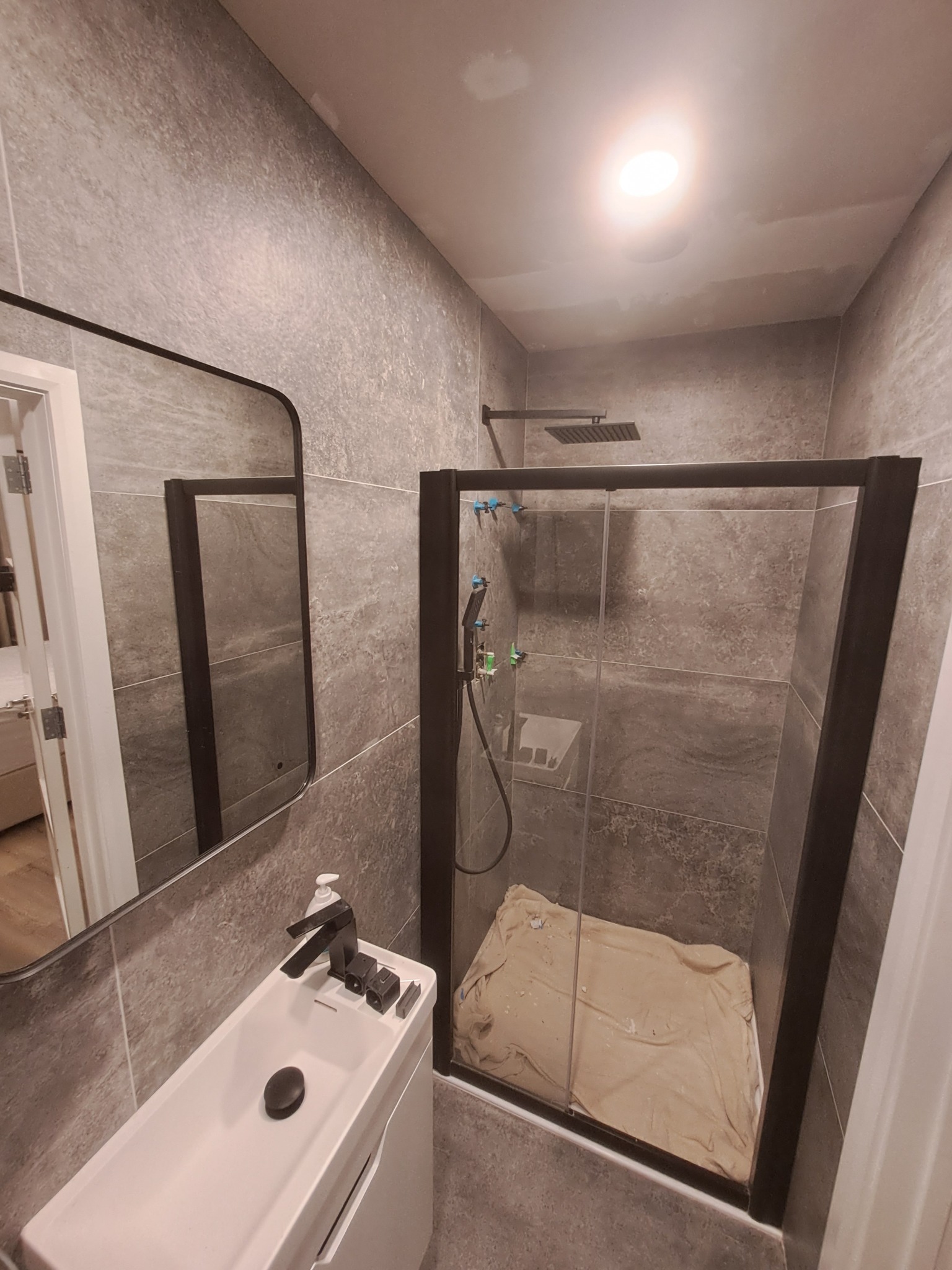 Nowoczesna łazienka w Nowym Sączu z szarymi płytkami, kabiną prysznicową z czarnymi ramami, minimalistyczną umywalką i lustrem. Pod prysznicem widoczne zabezpieczenie podłogi na czas prac...