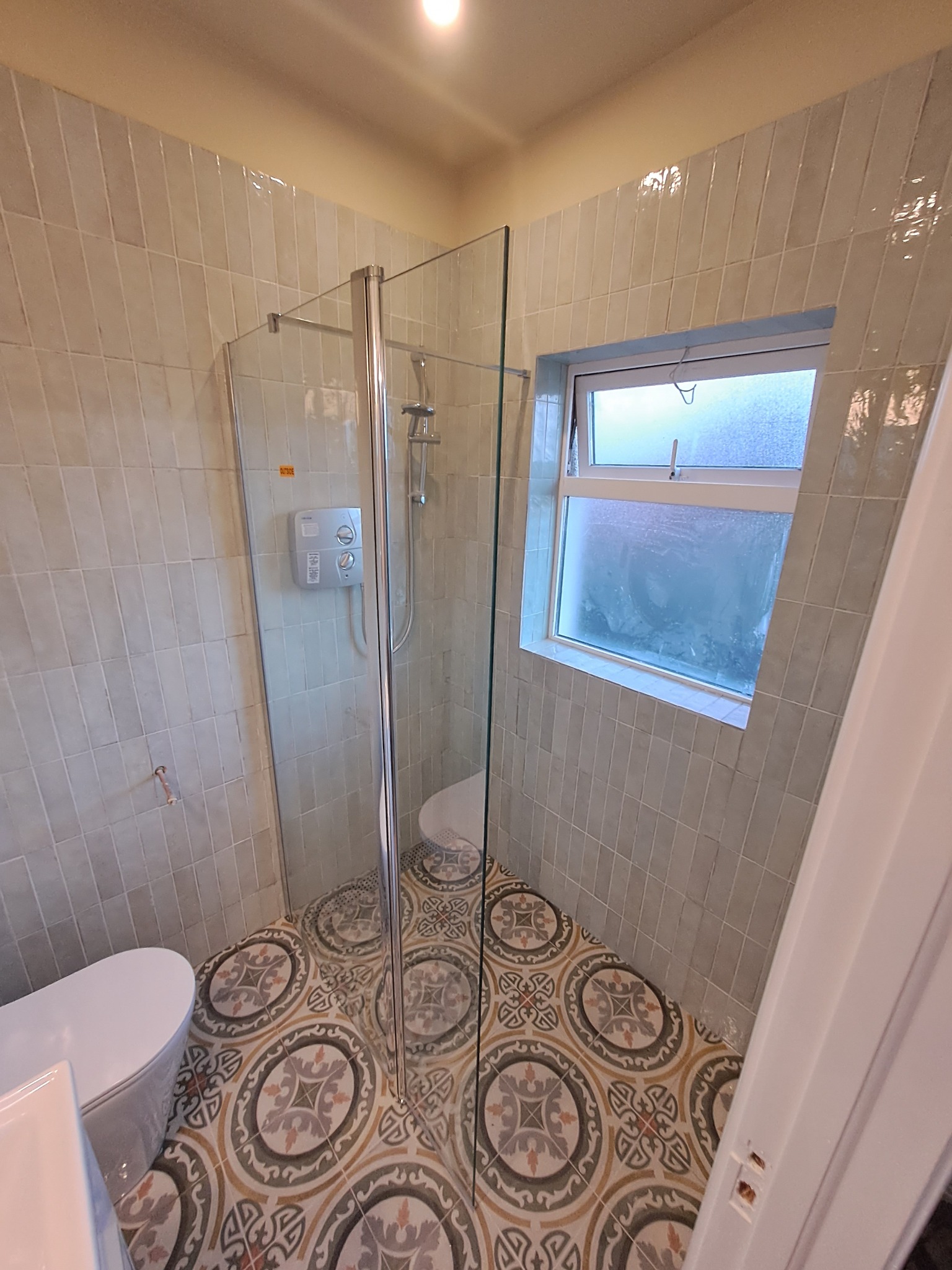 Nowoczesna łazienka po remoncie: kabina prysznicowa ze szklaną ścianką, ozdobne płytki podłogowe z motywem geometrycznym, ściany wyłożone pionowymi, jasnymi płytkami imitującymi cegłę, okno...