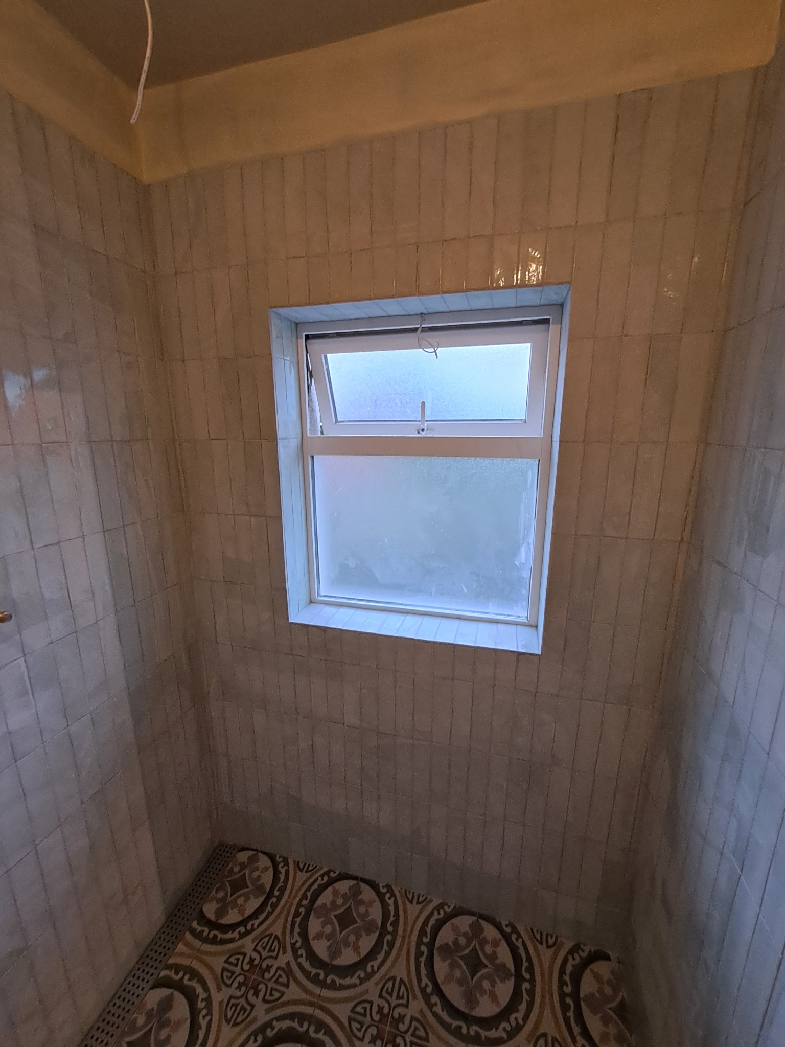 Wykończona łazienka z beżowymi, pionowo ułożonymi płytkami ściennymi, odpływem liniowym i wzorzystą podłogą w stylu retro, okno z widokiem na zewnątrz.