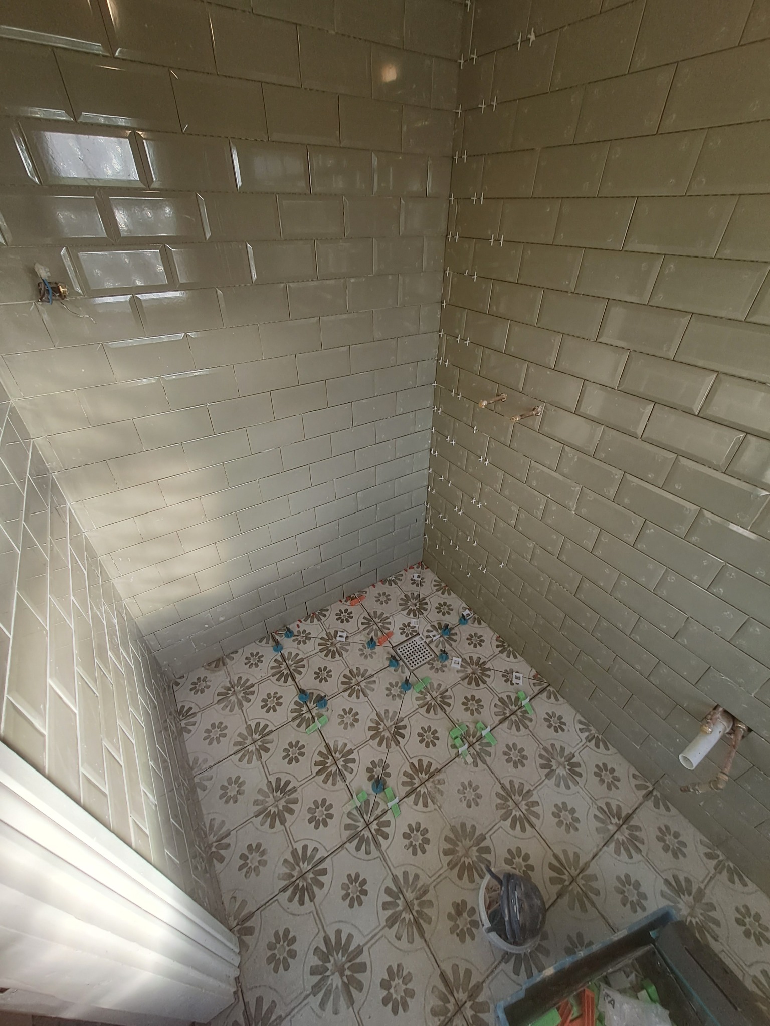 Wnętrze łazienki w trakcie remontu: połyskujące, oliwkowe płytki ścienne ułożone w cegiełkę z białymi krzyżykami dystansowymi, podłoga z wzorzystych płytek, widoczne rury i puszki elektryczne.