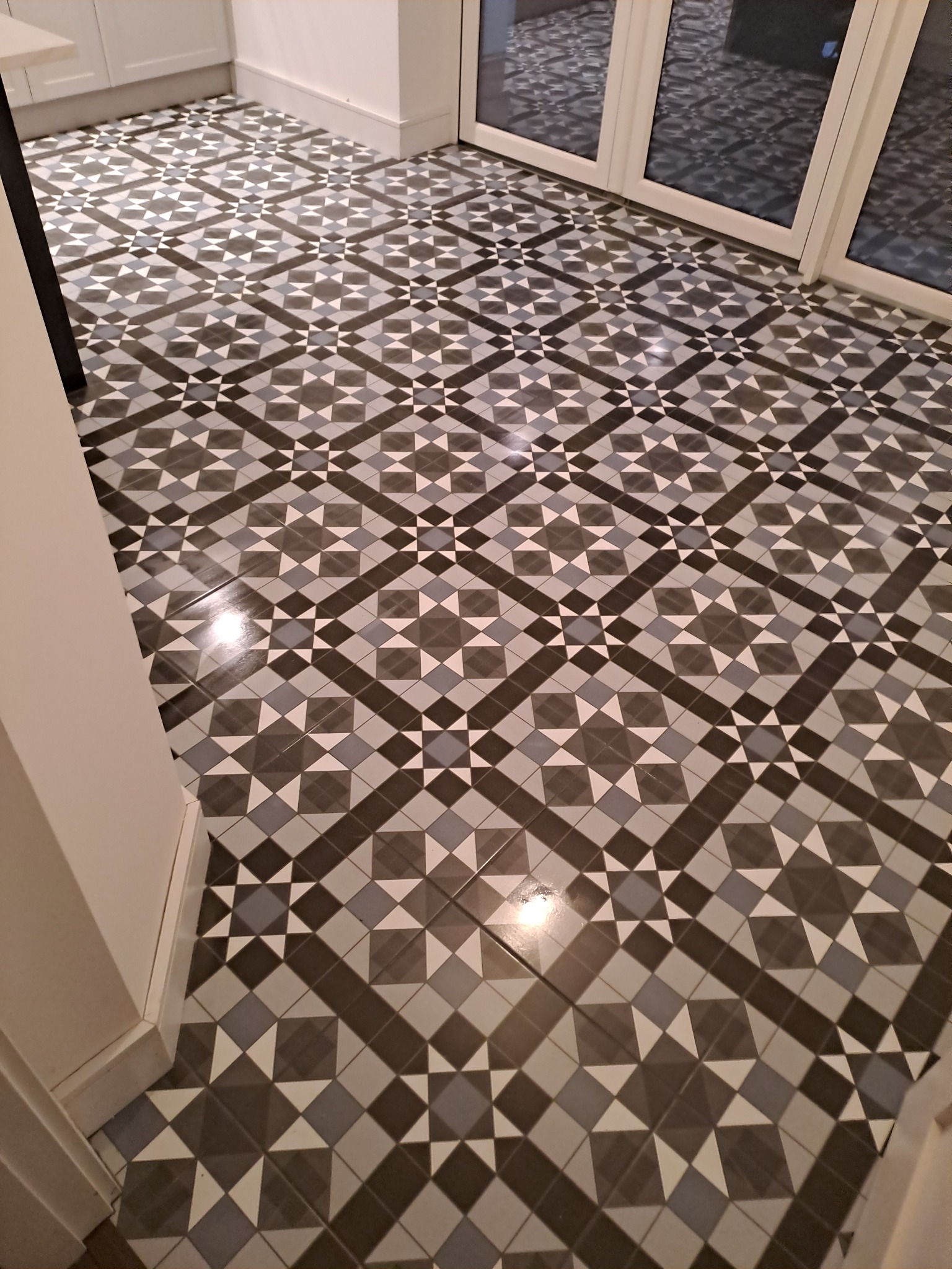 Wzorzysta podłoga z płytek ceramicznych w odcieniach szarości i bieli, ułożona w geometryczny, symetryczny deseń, widoczna w częściowo oświetlonym pomieszczeniu z białymi ścianami i drzwiami...