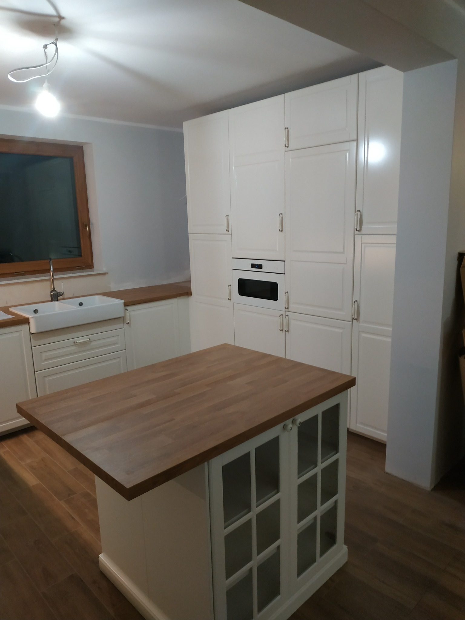 Wnętrze kuchni z białymi szafkami, drewnianym blatem i wyspą kuchenną z przeszklonymi drzwiczkami, oświetlone pojedynczą lampą wiszącą.