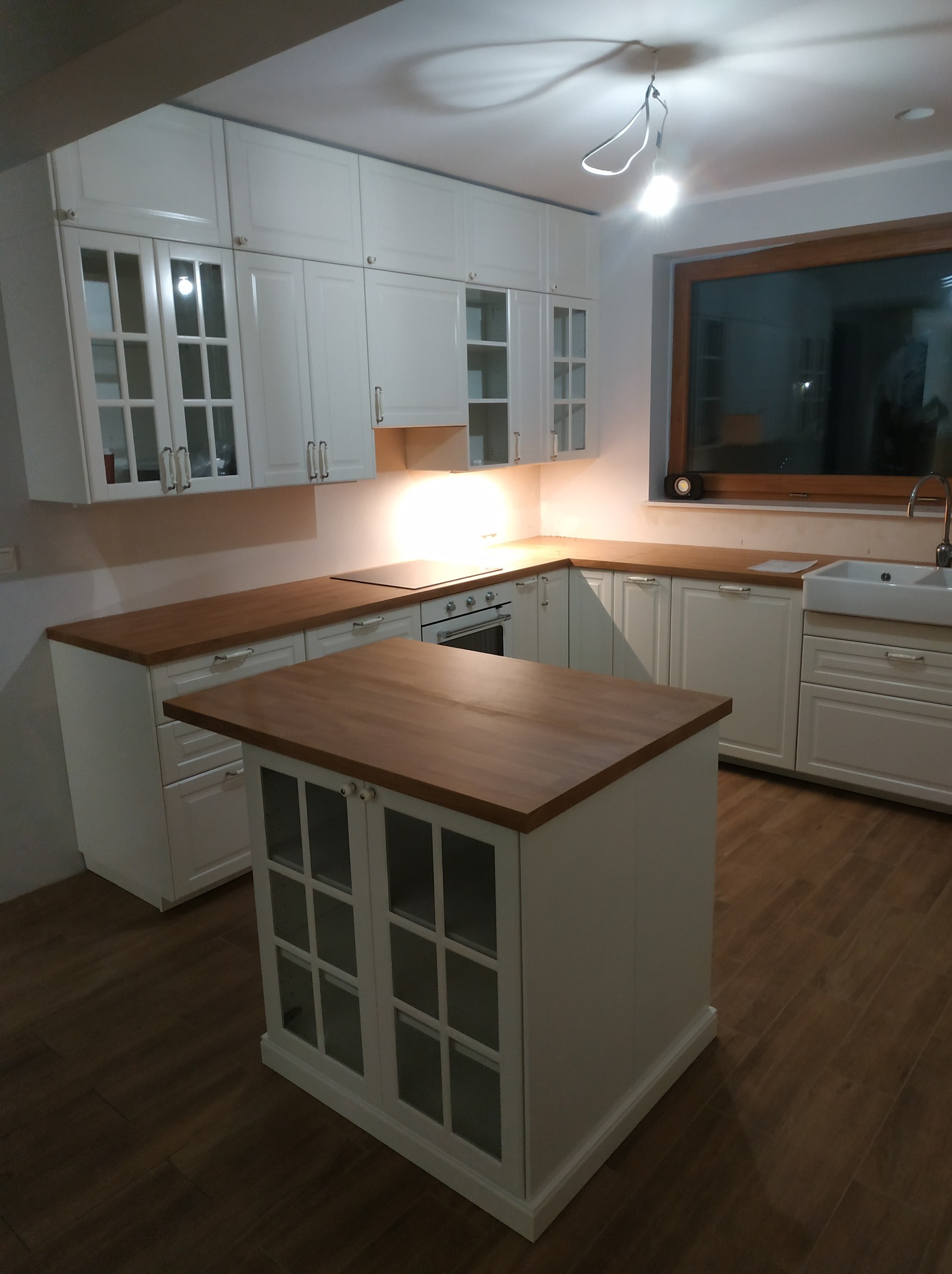 Jasna kuchnia z drewnianym blatem i wyspą kuchenną z przeszklonymi drzwiczkami, widok na zlewozmywak i okno.