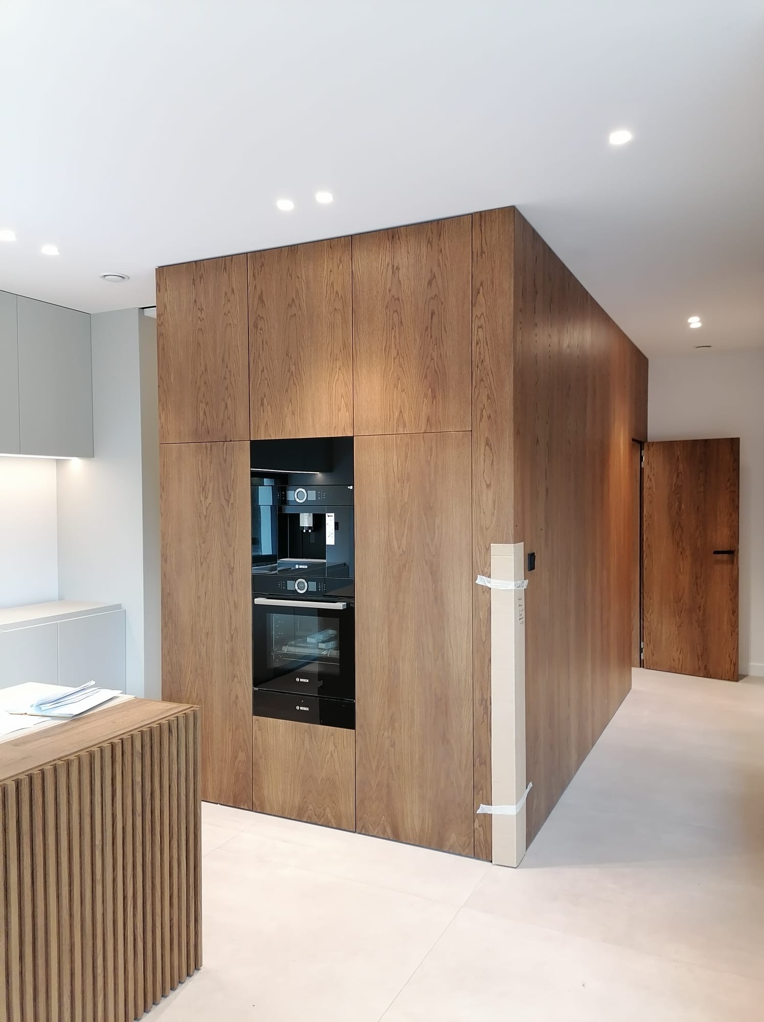 Nowoczesna kuchnia z zabudową z drewnianych paneli, w której wkomponowano piekarnik i ekspres do kawy marki Bosch. Widoczny fragment wyspy kuchennej z pionowymi listwami oraz niezapakowany karton...