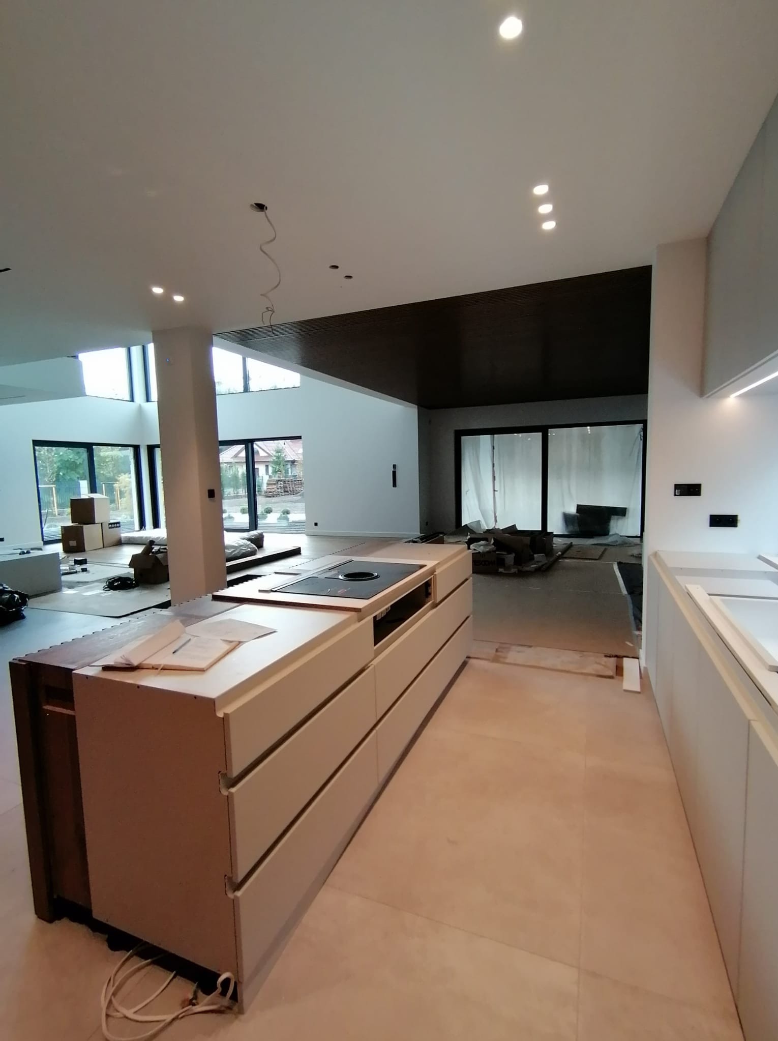 Wnętrze w trakcie montażu kuchni: wyspa kuchenna z płytą grzewczą, widoczne kable i elementy instalacji, podłoga w trakcie wykończenia, duże okna w tle.