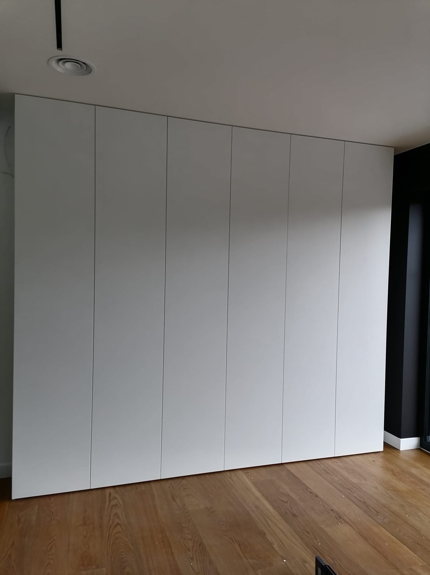 Biała szafa na wymiar z siedmioma drzwiami, parkiet dębowy na podłodze i czarna rama okienna po prawej stronie.