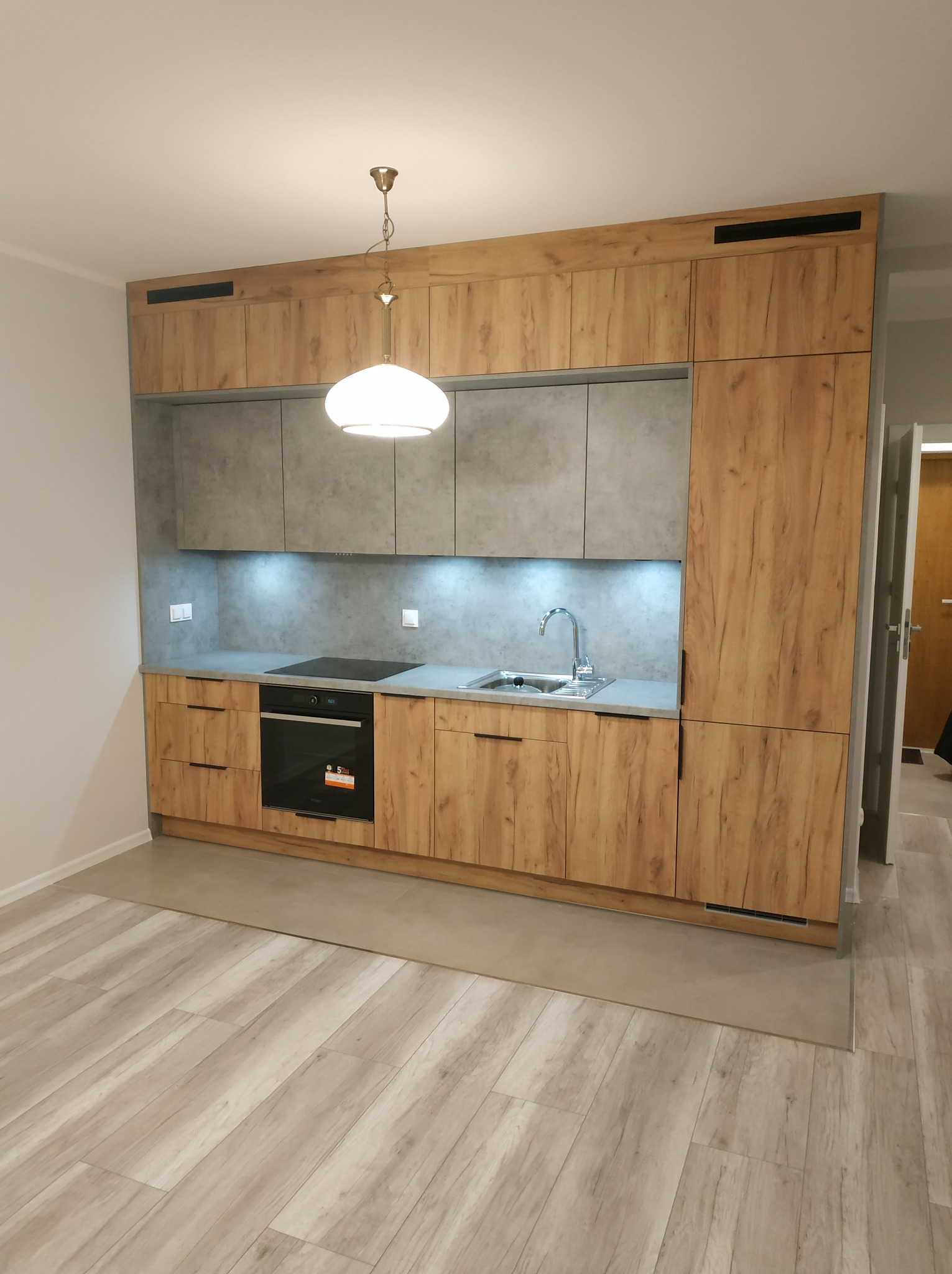 Nowoczesna kuchnia z drewnianymi frontami szafek i szarymi górnymi szafkami, oświetleniem LED pod szafkami, płytą indukcyjną i jasną podłogą.