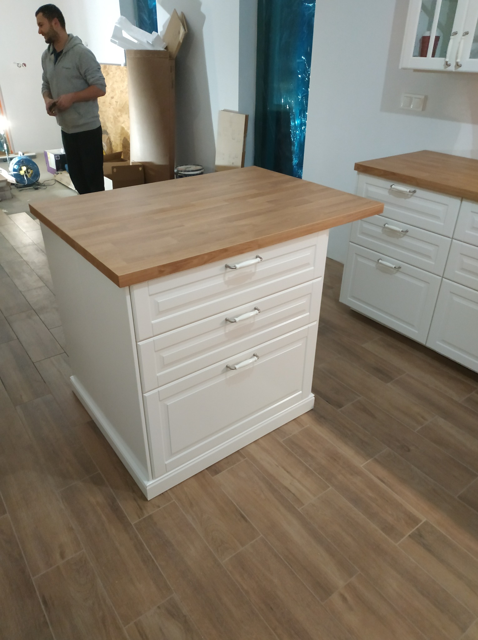 Wyspa kuchenna z trzema szufladami i drewnianym blatem, widoczna w trakcie montażu, z fragmentem zabudowy kuchennej w tle i osobą w ubraniu roboczym w tle.