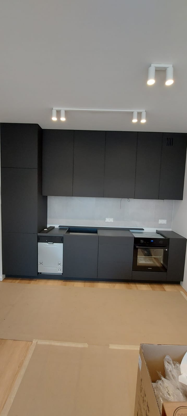 Nowoczesna, czarna zabudowa kuchenna z oświetleniem punktowym, widoczna z perspektywy podłogi zabezpieczonej kartonem, sugerująca etap montażu.