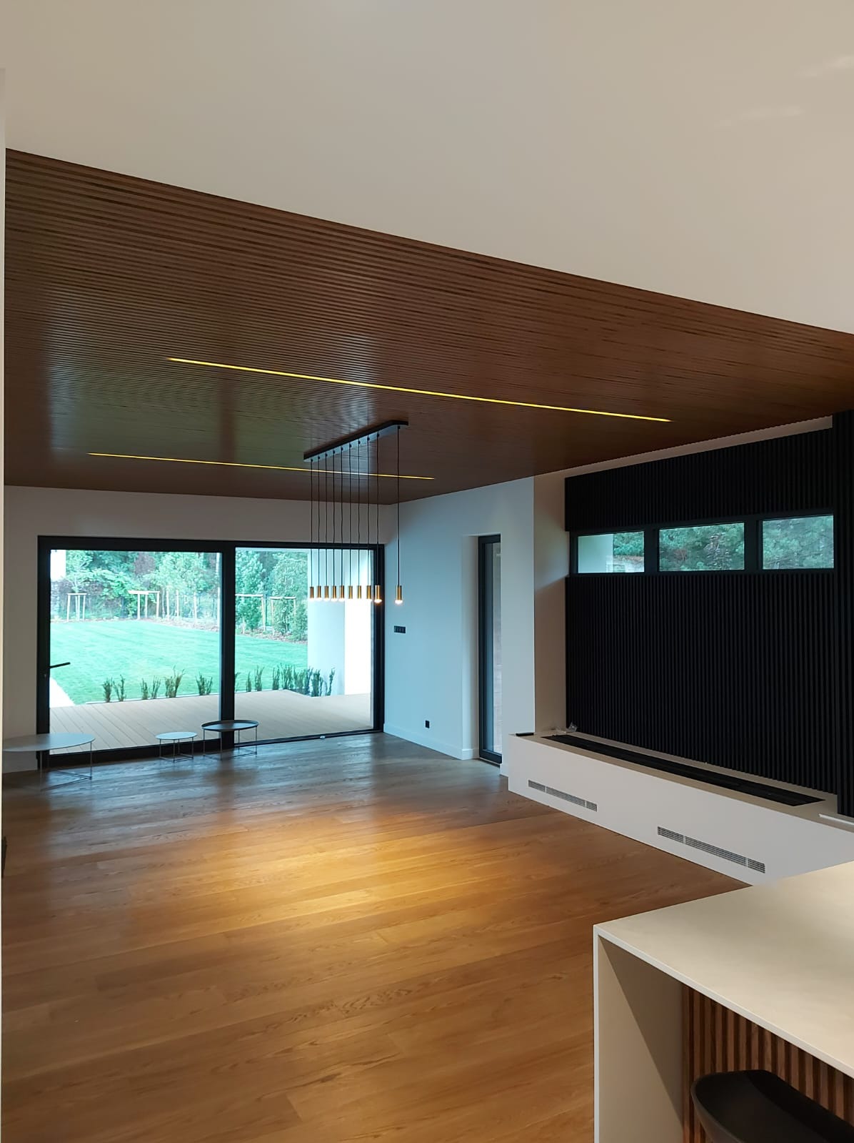 Nowoczesny salon z drewnianym sufitem lamelowym, podświetleniem LED, podłogą z jasnego drewna i czarną ścianą z pionowych paneli. Widoczne przesuwne okno z wyjściem na taras i ogród.