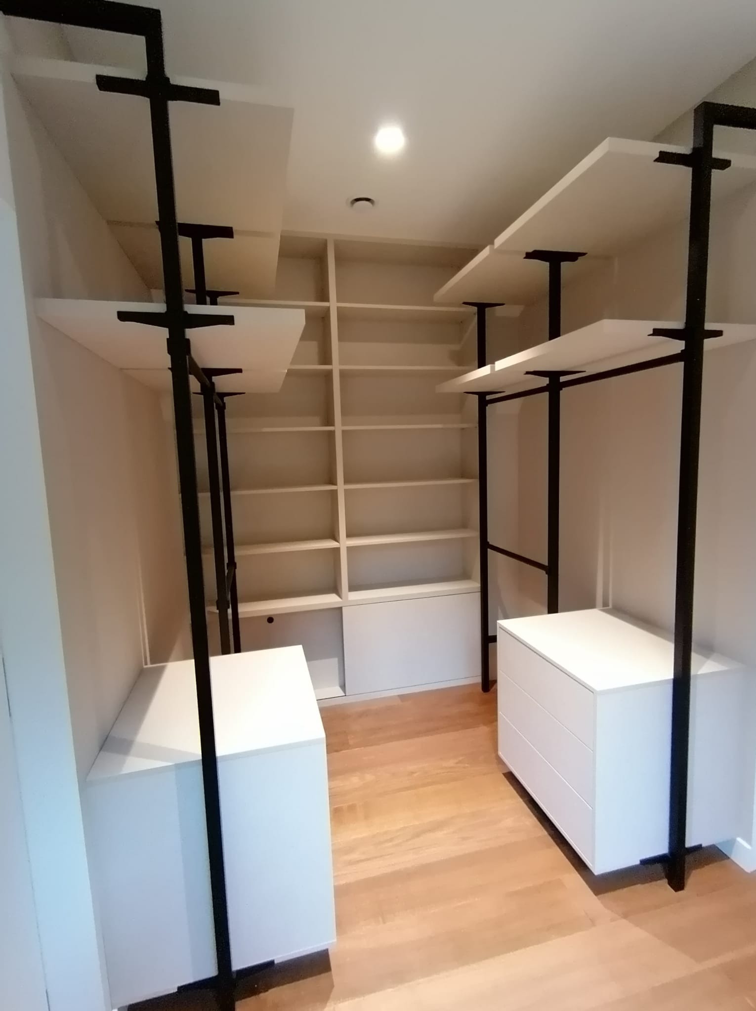 Wnętrze garderoby z białymi szafkami i półkami na czarnych metalowych stelażach, podłoga z jasnego drewna, w tle regał z półkami.