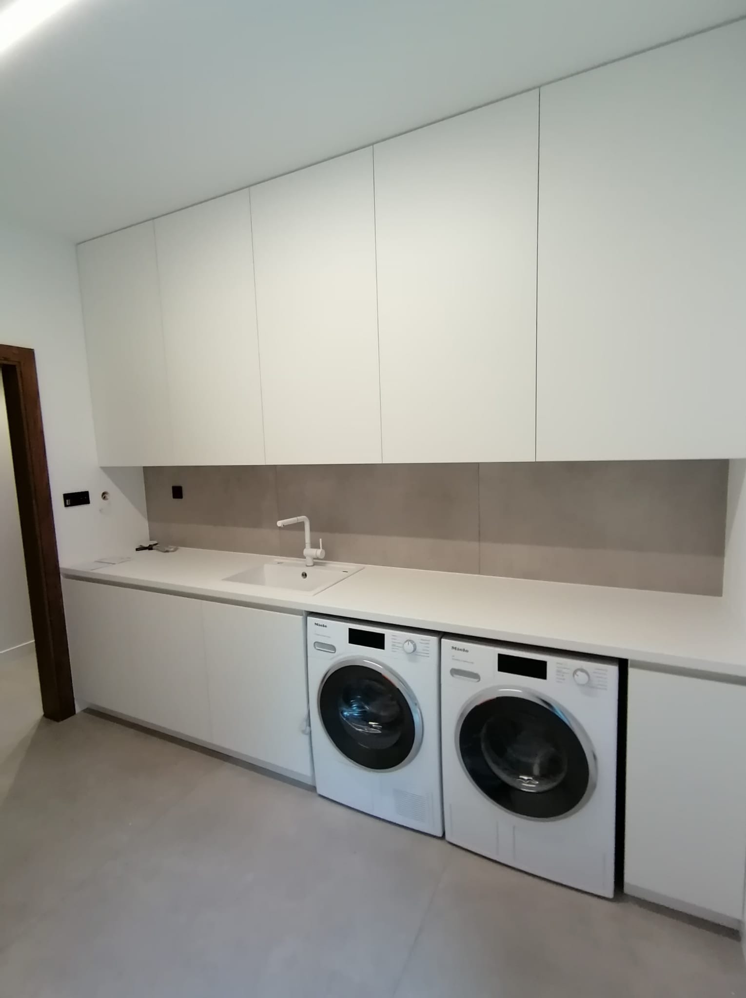 Nowoczesna, minimalistyczna pralnia z białymi szafkami na wymiar, blatem z wbudowanym zlewem, dwoma pralkami Miele i szarą ścianą w tle.