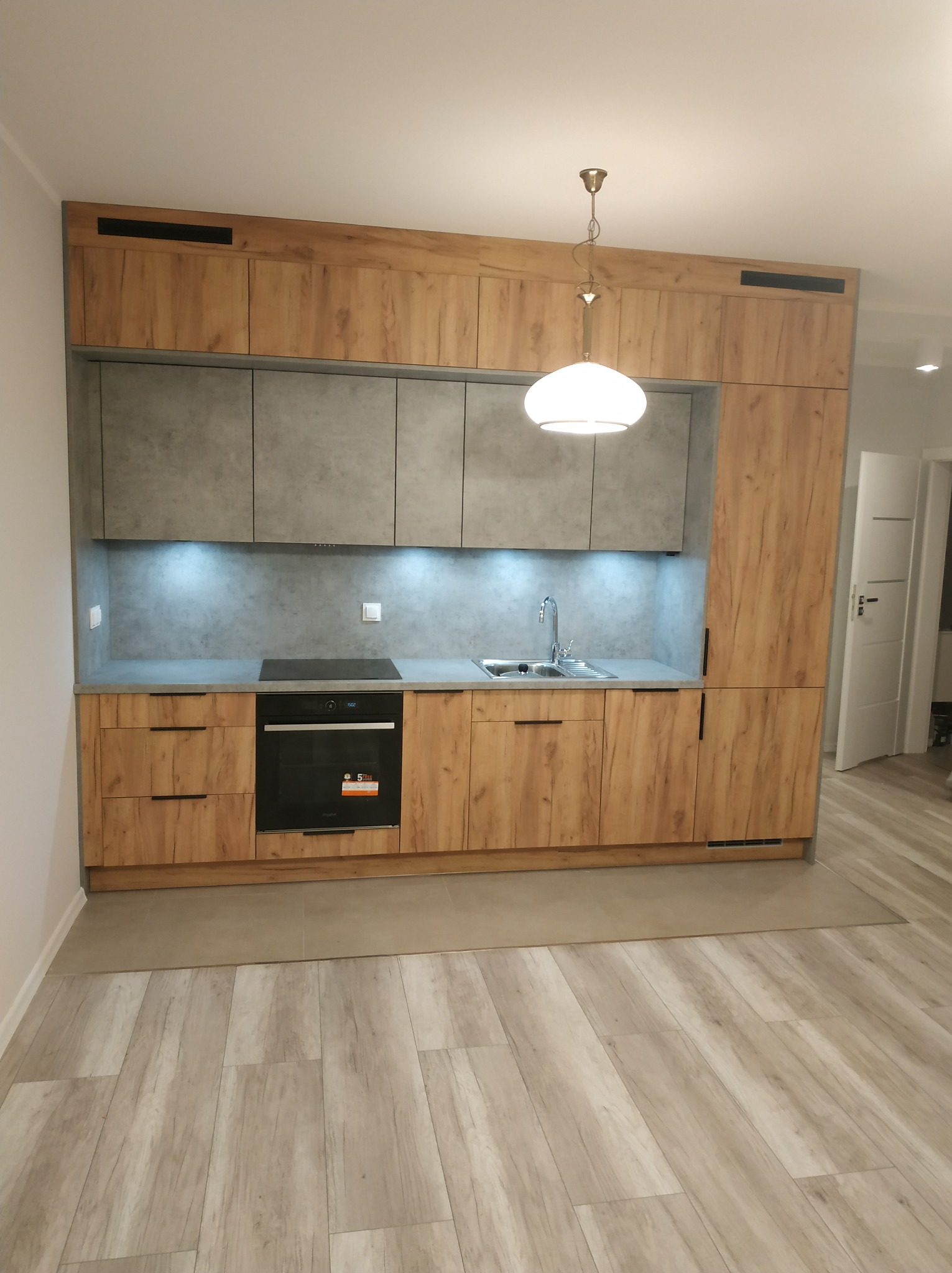 Nowoczesna kuchnia z drewnopodobnymi frontami szafek i szarym blatem, oświetlenie LED pod szafkami wiszącymi, wisząca lampa z okrągłym kloszem, płyta indukcyjna i piekarnik w zabudowie.