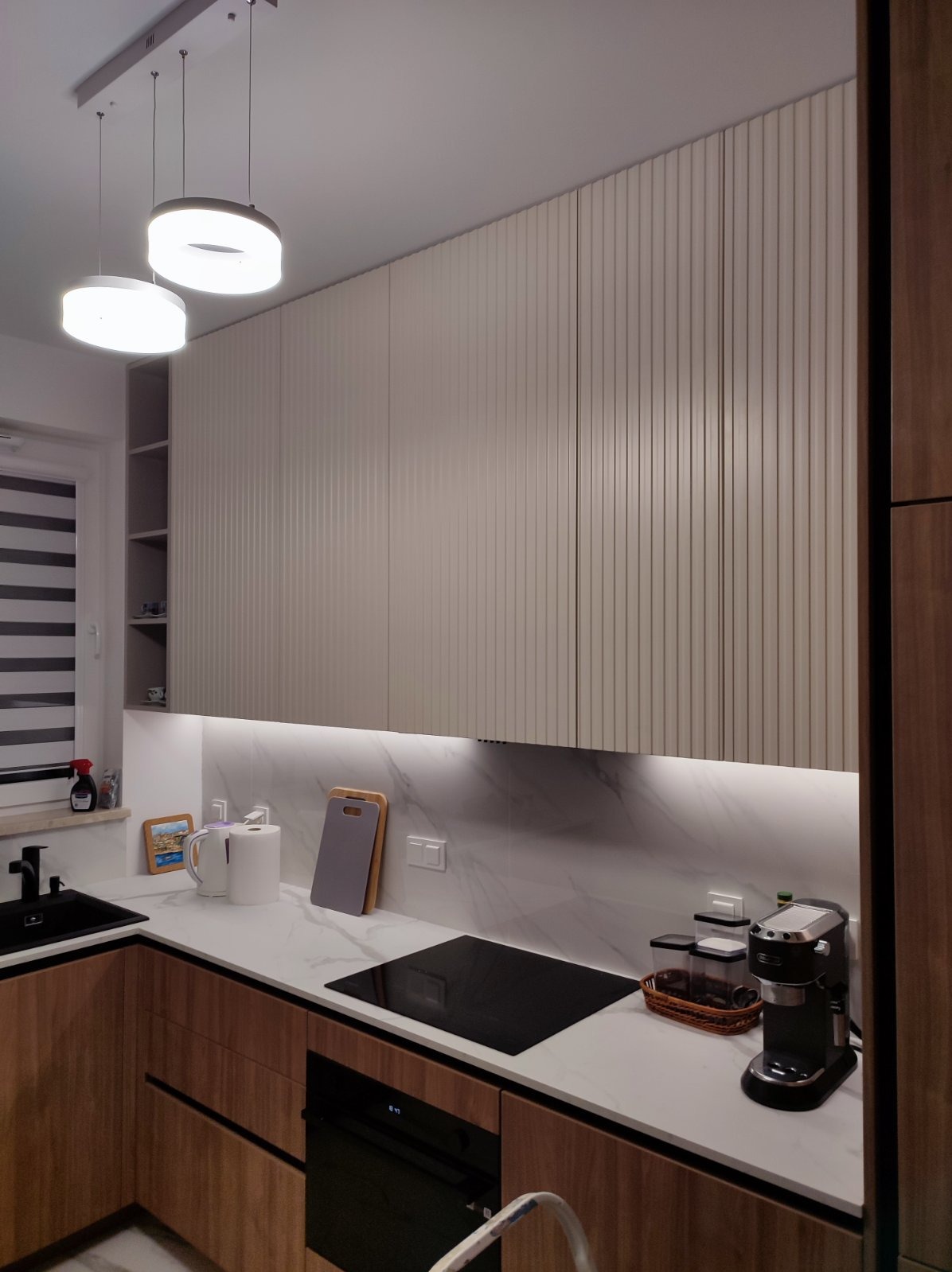 Nowoczesna kuchnia z frezowanymi frontami szafek górnych w kolorze beżowym, podświetleniem LED pod szafkami i blatem z białego marmuru, widoczny czarny zlew, płyta indukcyjna i ekspres do kawy.