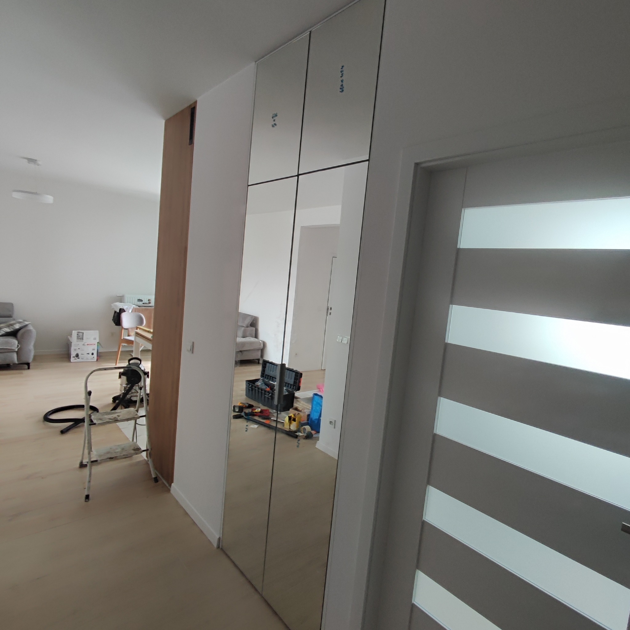 Montaż szafy z lustrzanymi drzwiami przesuwnymi w nowoczesnym mieszkaniu, widoczna skrzynka z narzędziami, drabina i odkurzacz, częściowo odbicie wnętrza pokoju.