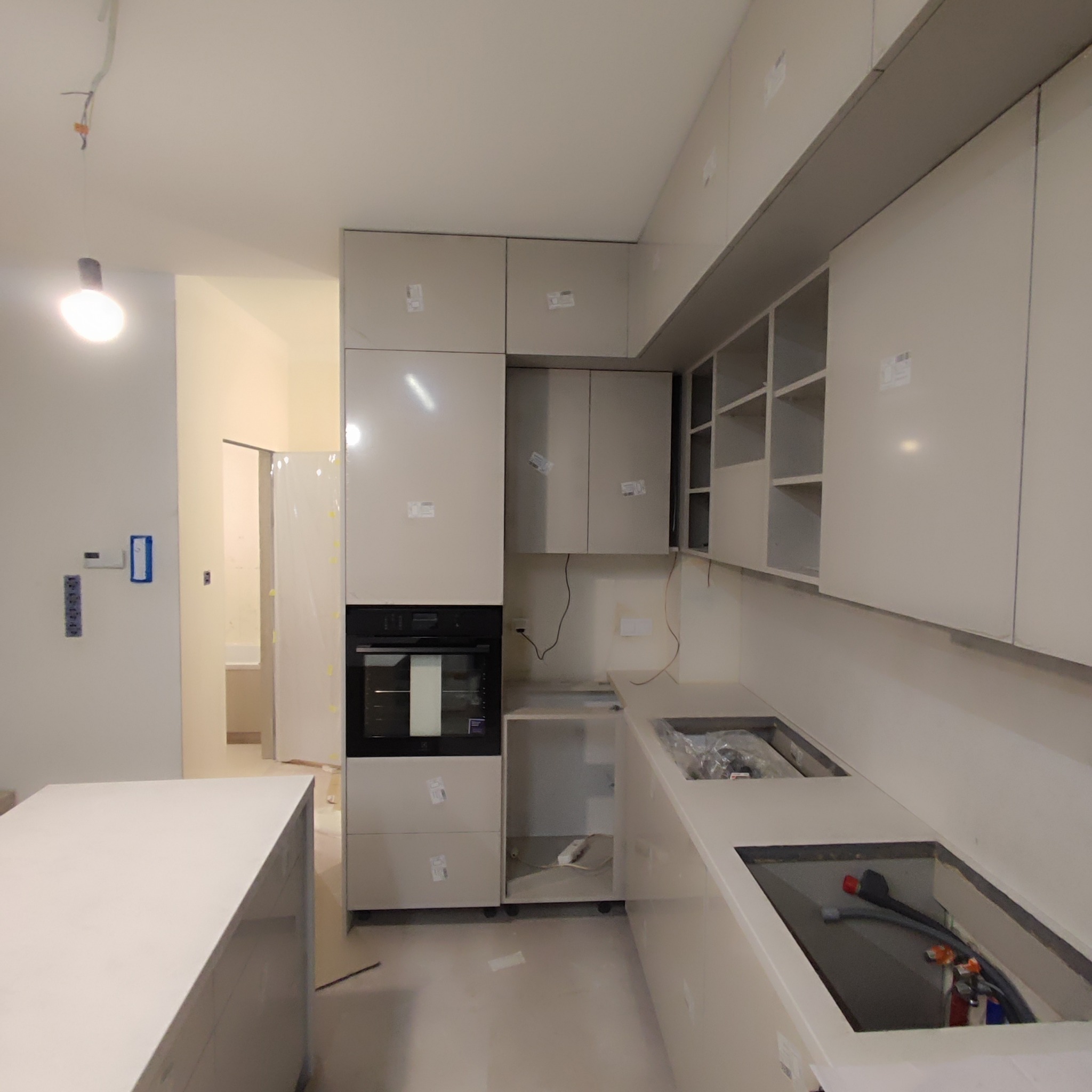 Wnętrze kuchni w trakcie montażu, widoczne białe szafki bez uchwytów, wbudowany piekarnik, wyspa kuchenna i niezabezpieczone otwory na zlewozmywaki z widocznymi instalacjami hydraulicznymi.