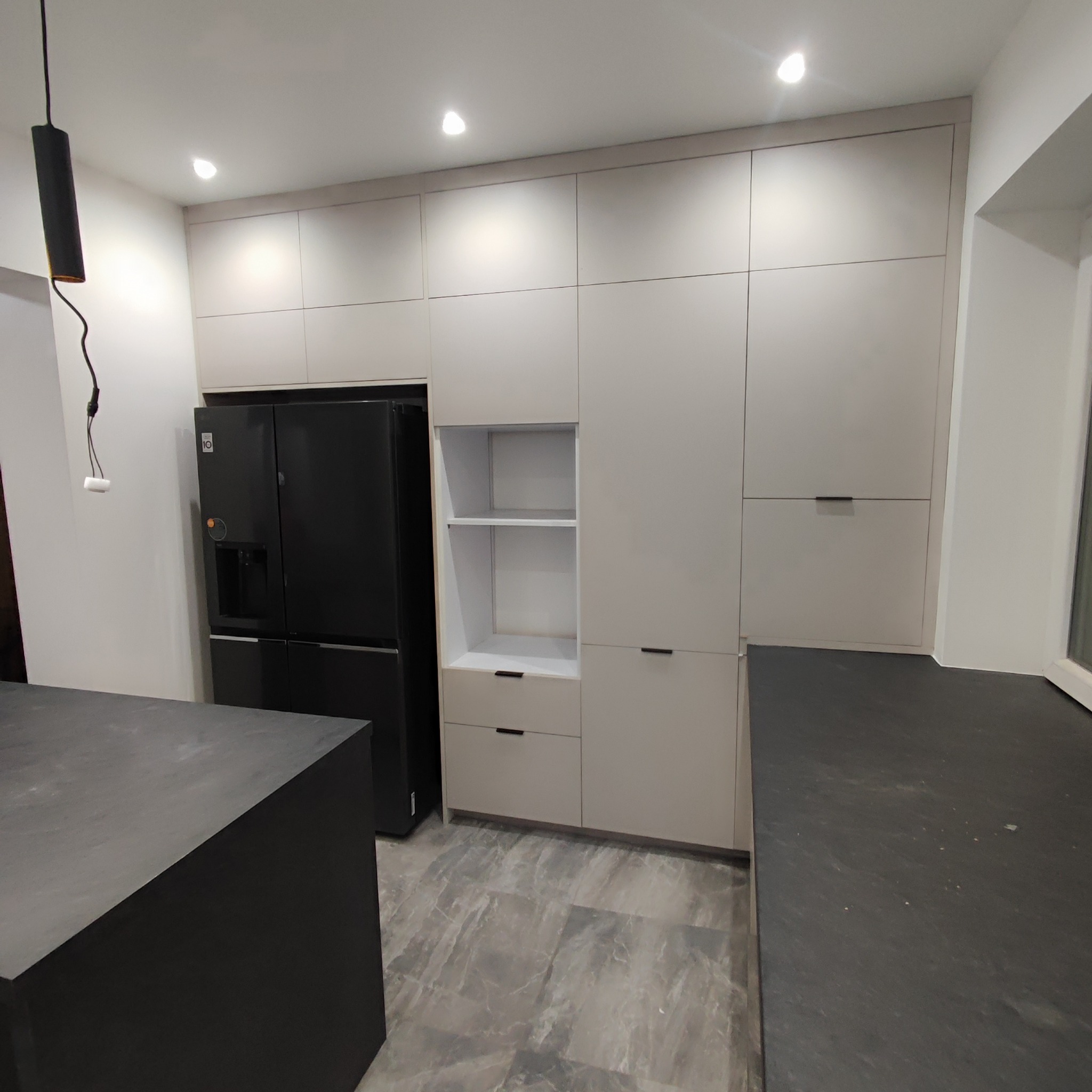 Nowoczesna kuchnia z zabudową w odcieniach szarości, czarna lodówka side-by-side, wyspa kuchenna z czarnym blatem, minimalistyczne uchwyty szafek.