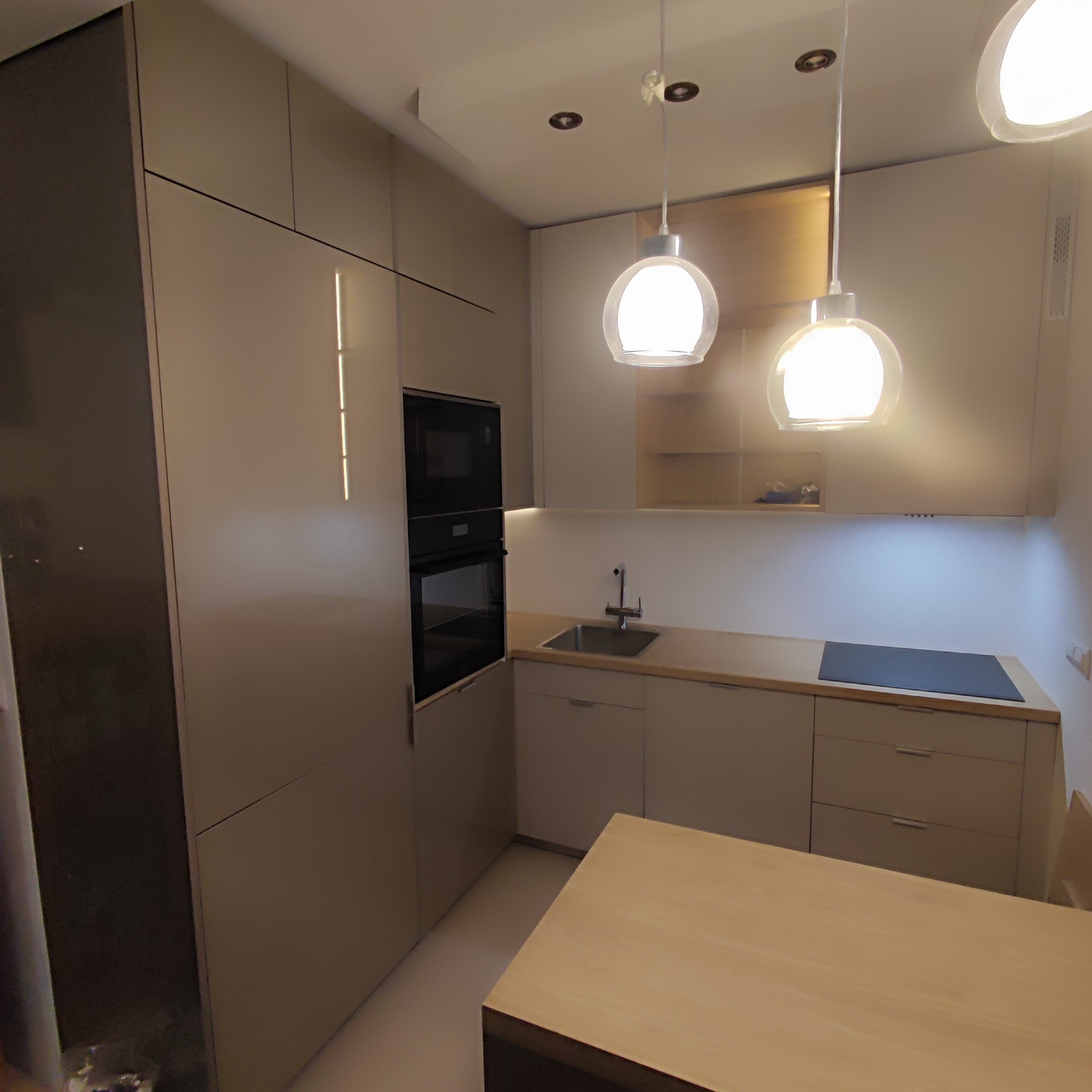 Jasna, nowoczesna kuchnia z zabudową pod sufit, oświetleniem punktowym i wiszącymi lampami kulami, z widocznym blatem roboczym, zlewem i płytą indukcyjną.