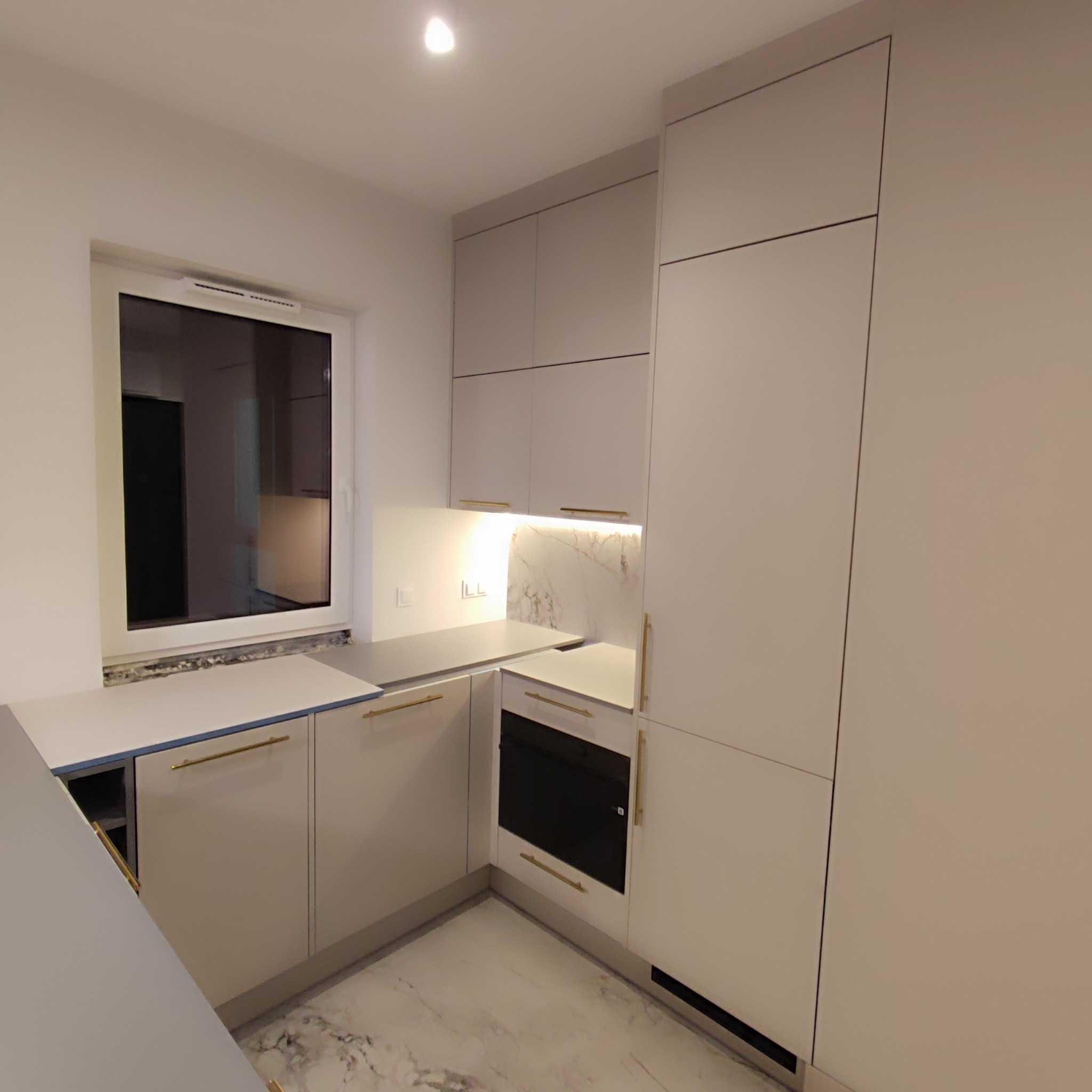 Jasna kuchnia na wymiar z marmurowym blatem i złotymi uchwytami, widok na okno i czarny piekarnik.