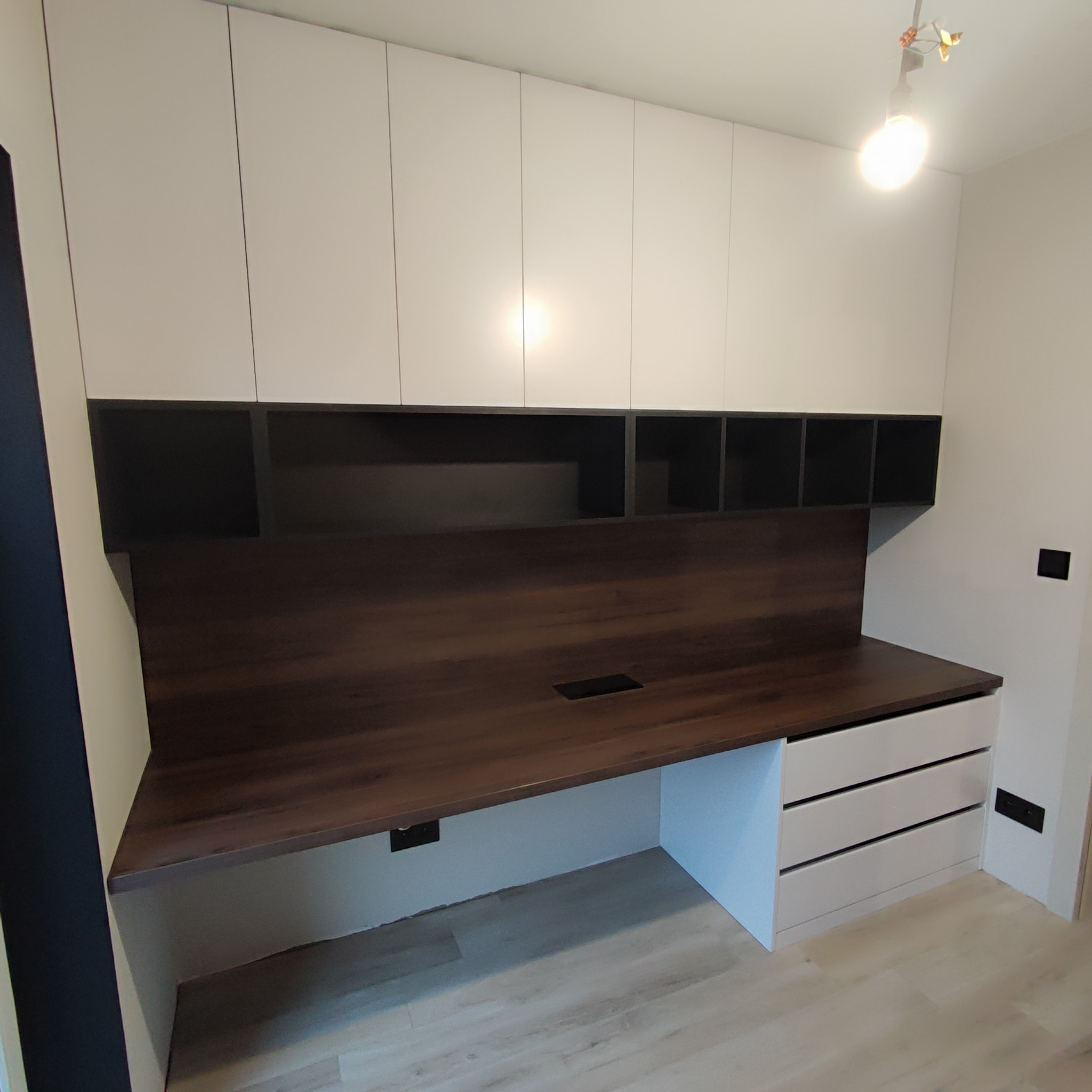 Biurko z blatem w kolorze ciemnego drewna, zintegrowane z białymi szafkami wiszącymi i szufladami, gniazdko elektryczne widoczne pod blatem, minimalistyczny design.
