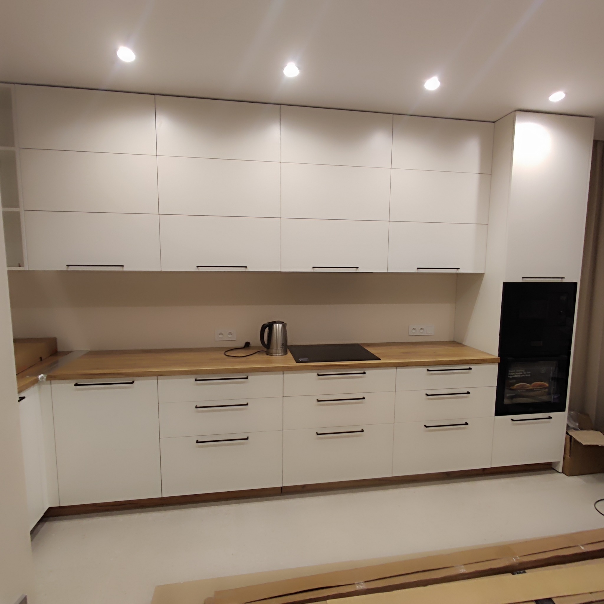 Nowoczesna kuchnia w bieli z drewnianym blatem, czarnymi uchwytami i wbudowanym piekarnikiem, widoczne kartony na podłodze sugerują niedawny montaż.