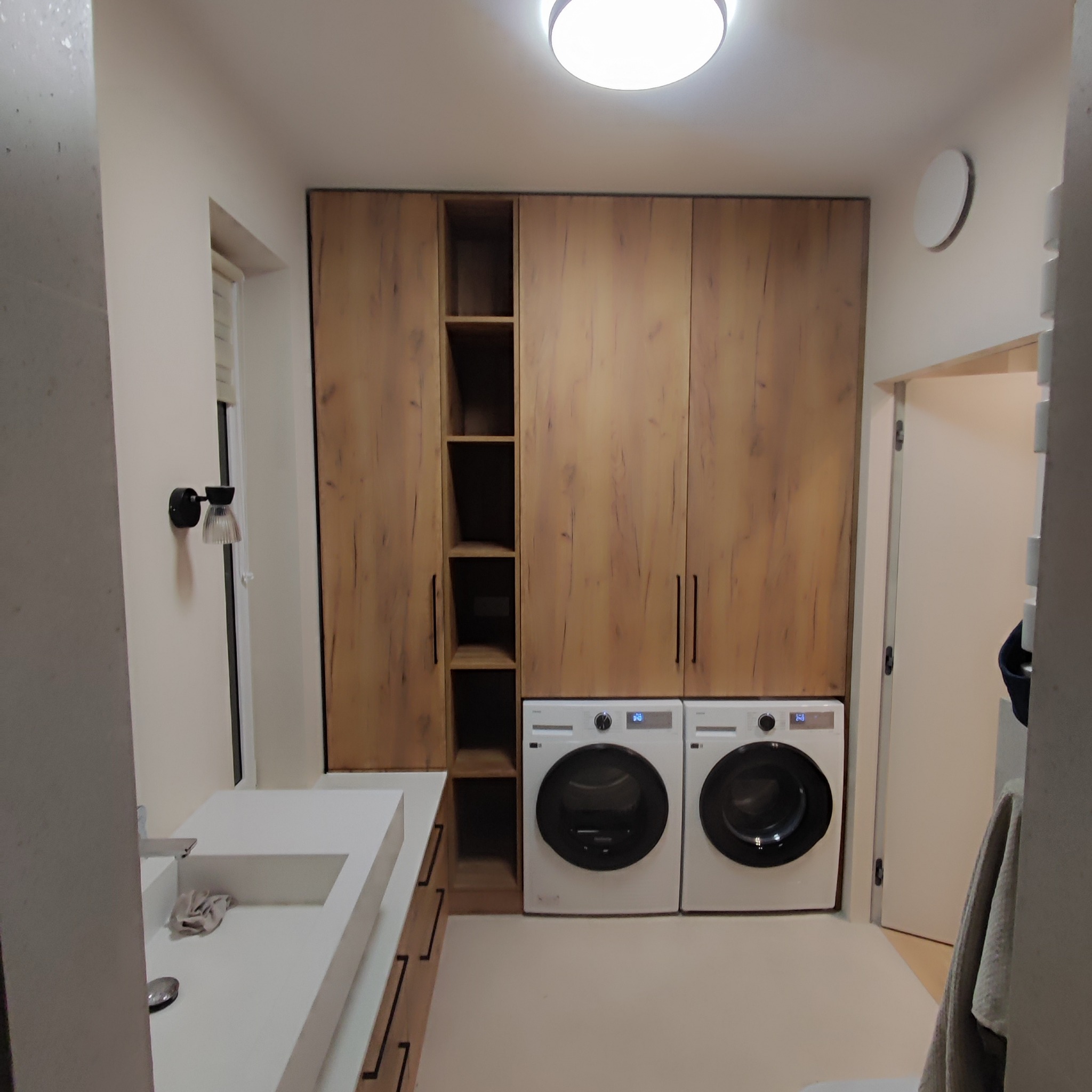 Pralka i suszarka w zabudowie z szafami z drewnopodobnymi frontami, minimalistyczna umywalka nablatowa, oświetlenie sufitowe i ścienne w pomieszczeniu gospodarczym.