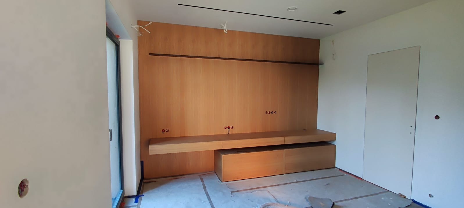 Wnętrze pokoju z drewnianą zabudową ścienną, wiszącą szafką RTV i ukrytymi kablami elektrycznymi, widoczne ślady prac wykończeniowych na podłodze.