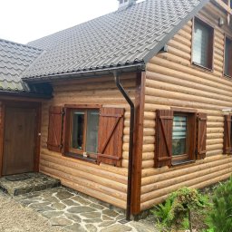 Artur Wróblewski Usługi remontowo-budowlane - Zewnętrzny widok domu z bali z drewnianymi okiennicami i kamiennymi schodami wejściowymi.