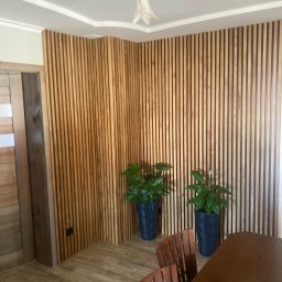 Artur Wróblewski Usługi remontowo-budowlane - Drewniane lamele pionowe na ścianie w salonie, dwie donice z roślinami, widoczny fragment stołu i krzeseł oraz lampa sufitowa w kształcie kwiatów.
