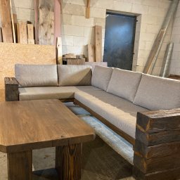 Artur Wróblewski Usługi remontowo-budowlane - Kanciasty, drewniany stół i sofa narożna z jasną tapicerką, z grubych belek, w warsztacie z betonowych pustaków.