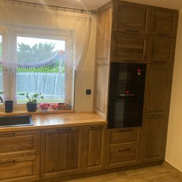 Artur Wróblewski Usługi remontowo-budowlane - Kuchenna zabudowa z drewnianymi szafkami, blatem i wbudowanym piekarnikiem, z czarnym zlewem pod oknem ozdobionym firanką i roślinami.