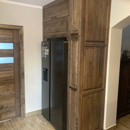 Artur Wróblewski Usługi remontowo-budowlane - Drewniana zabudowa lodówki w kuchni z drewnianymi drzwiami w tle, widoczne magnesy na lodówce.