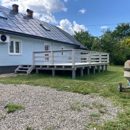 Artur Wróblewski Usługi remontowo-budowlane - Jasny dom z nowym, podwyższonym tarasem z drewnianą balustradą. Widoczne schody wejściowe i betonowa mieszarka. Błękitne niebo z obłokami w tle.