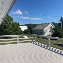 Artur Wróblewski Usługi remontowo-budowlane - Jasny, drewniany taras z barierkami w odcieniach szarości, widok na zielony ogród i dom w tle, słoneczny dzień, cień barierki na deskach.