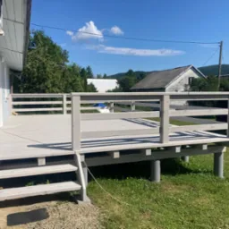 Jasnoszary taras z balustradą, widok na ogród i domy w tle. Schody prowadzą na taras. W rogu wkrętarka i pudełko z wkrętami.