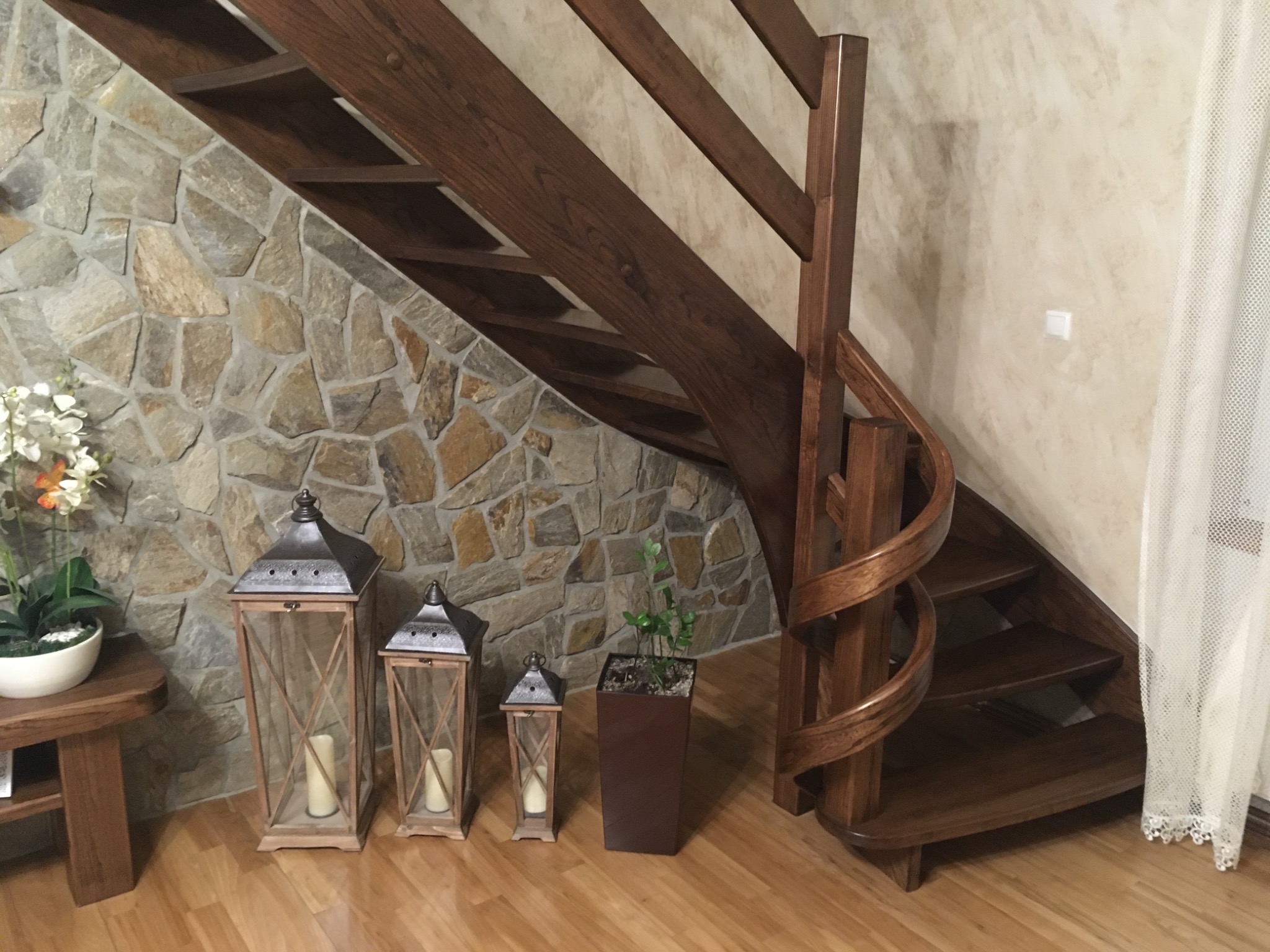 Drewniane schody kręte z ciemnego drewna, z kamienną ścianą w tle i dekoracyjnymi lampionami oraz roślinami doniczkowymi, oświetlone ciepłym światłem.